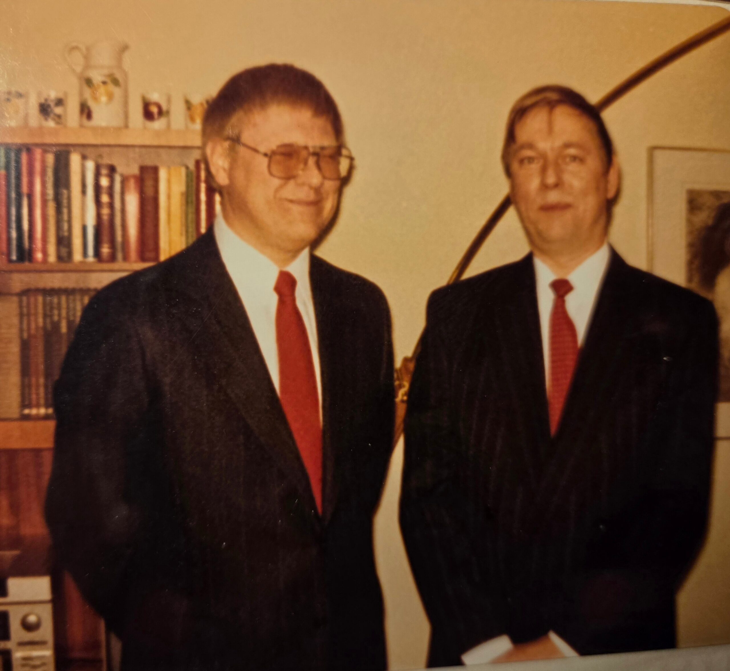 Two men in dark suits and red ties posing for a photo in front of a bookshelf in a study room.
