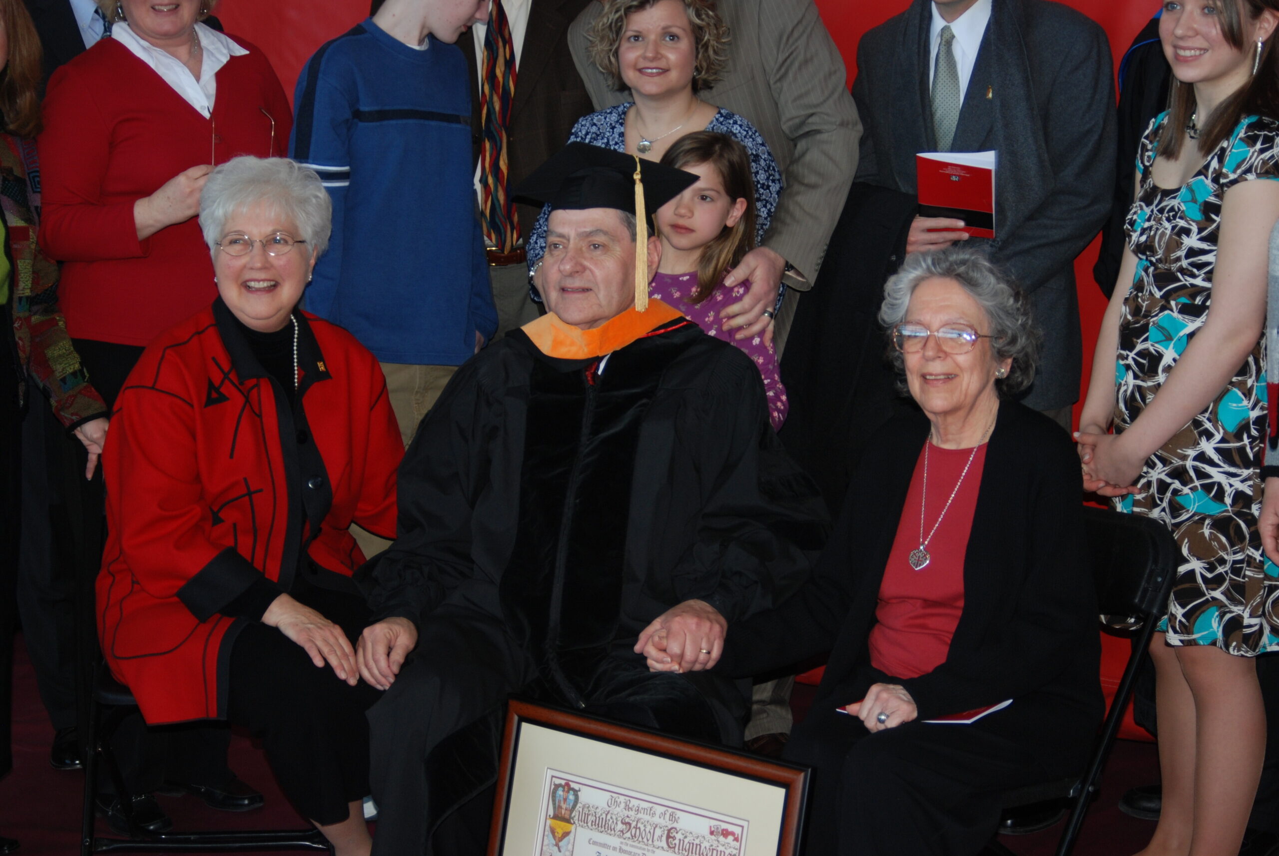 Graduate in cap and gown seated among smiling family and friends at a ceremony, with a framed certificate in the foreground.