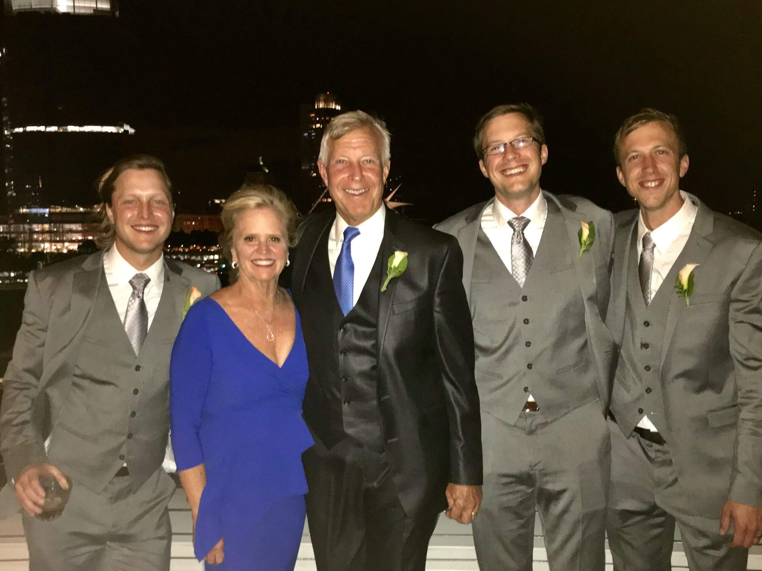Group of five people dressed in formal attire posing together at night, with four men in gray suits and a woman in a blue dress smiling at the camera.