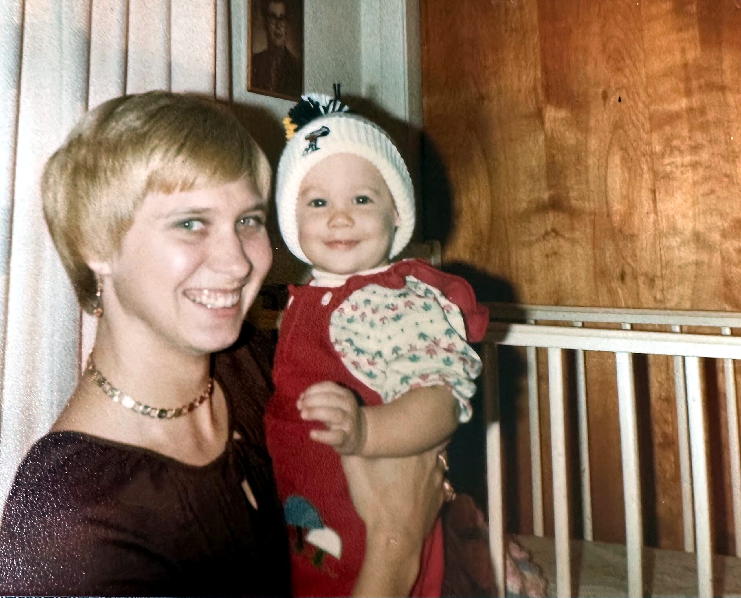 Smiling woman holding a baby in a knit hat indoors, both looking at the camera.