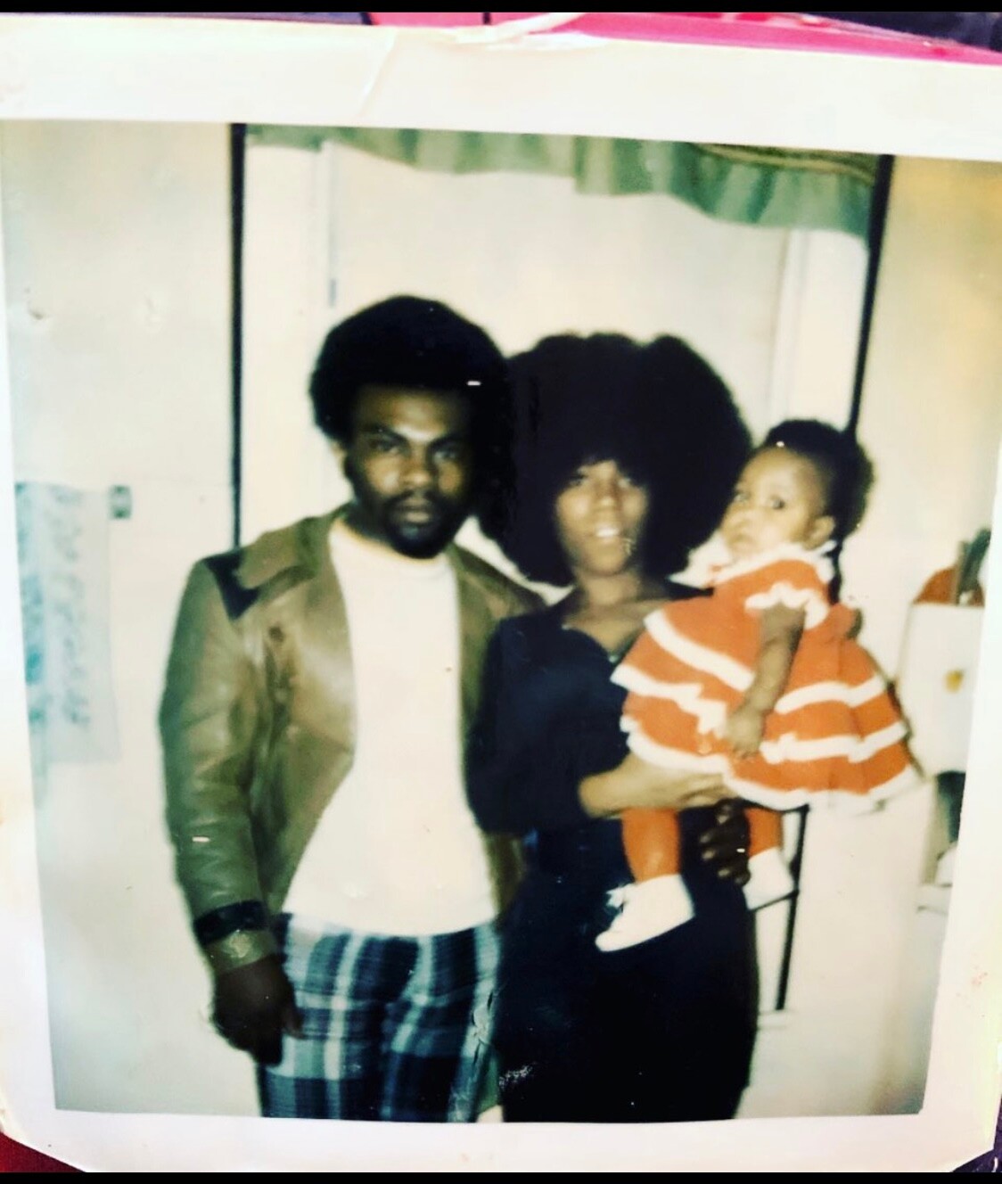 Family of three indoors: man in a tan jacket, woman with a large Afro holding a baby in orange stripes.