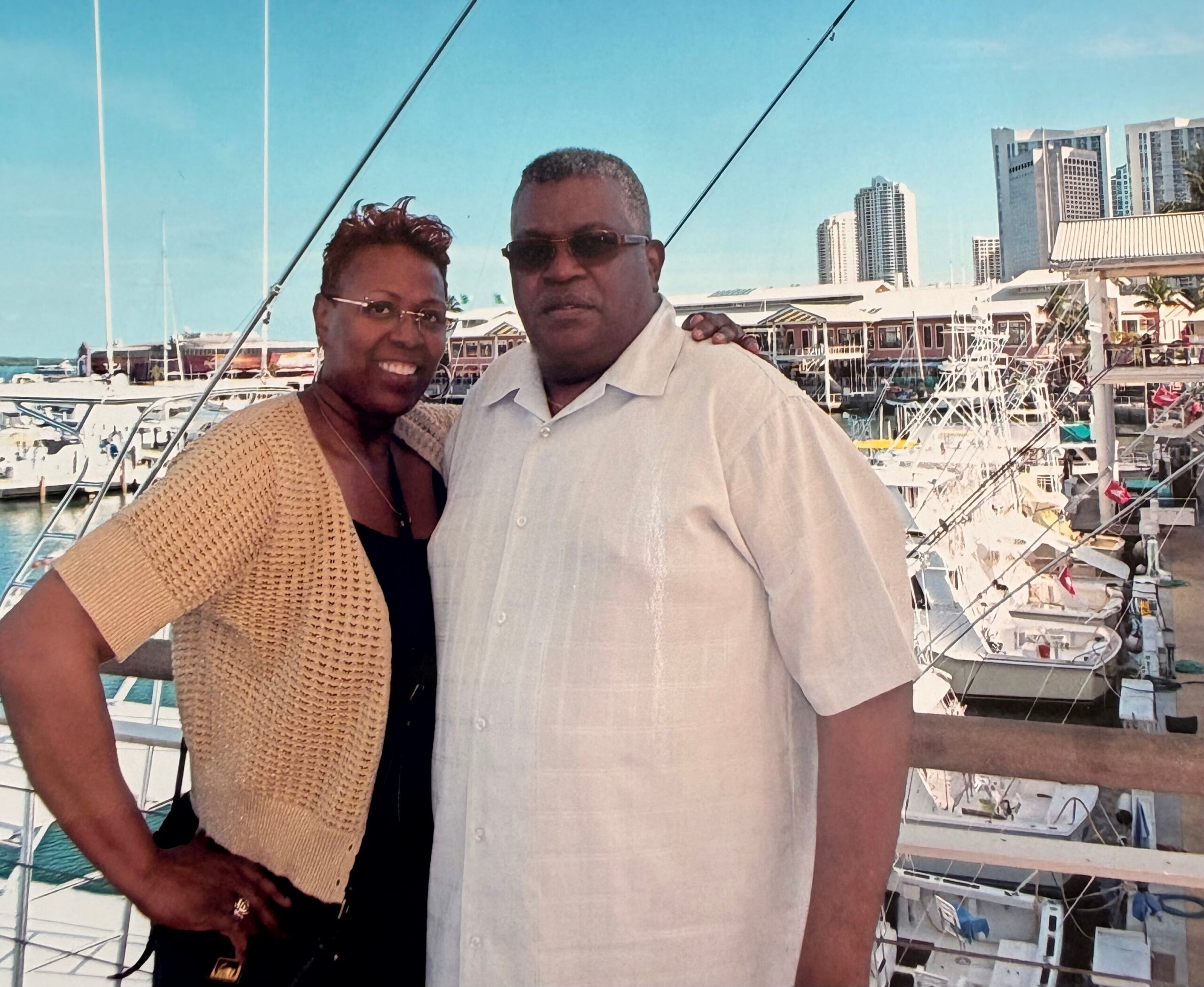 Smiling couple standing on a marina dock with sailboats and a city skyline in the background.