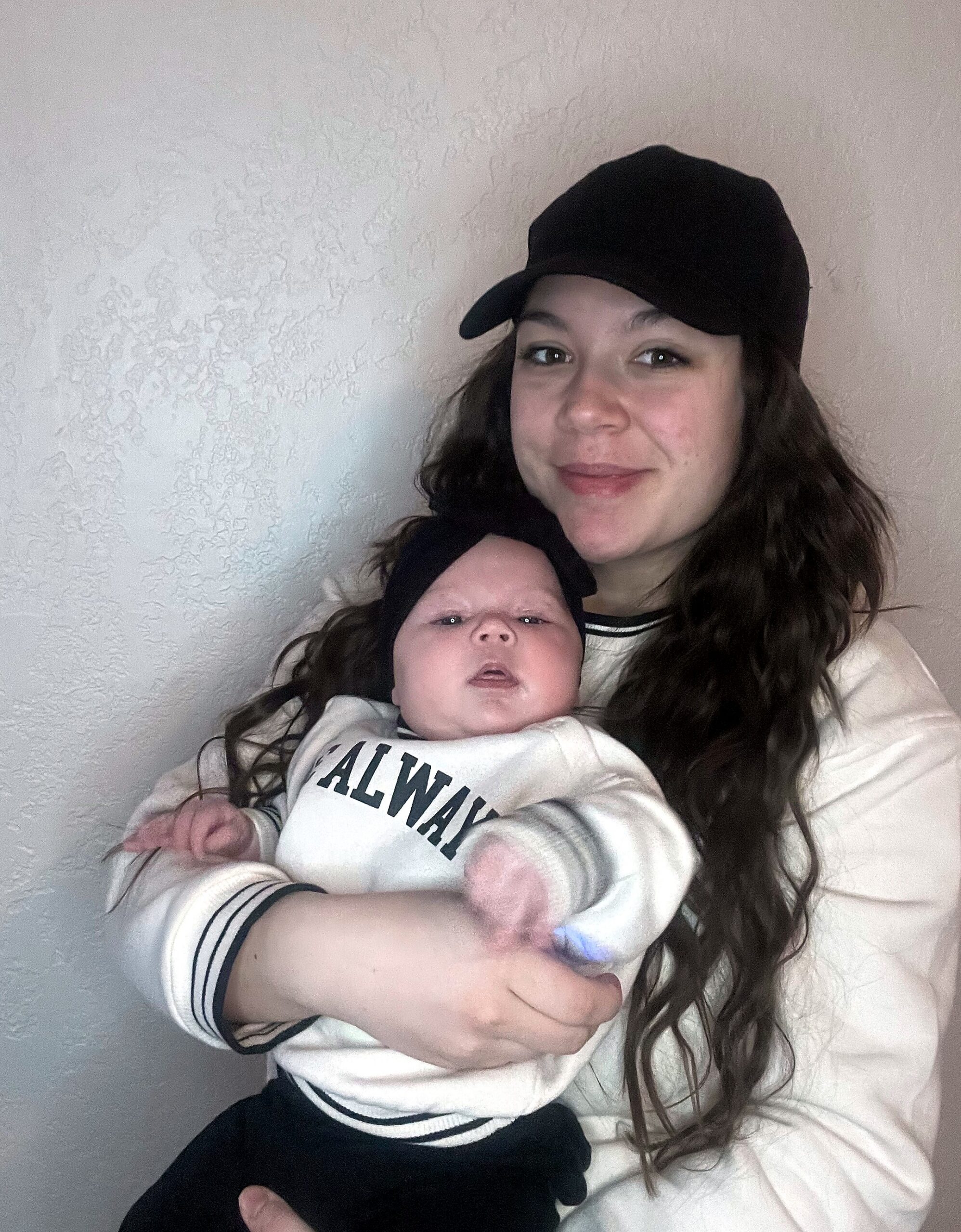 Woman wearing a black cap cradling a newborn baby in a white outfit, against a light textured wall.