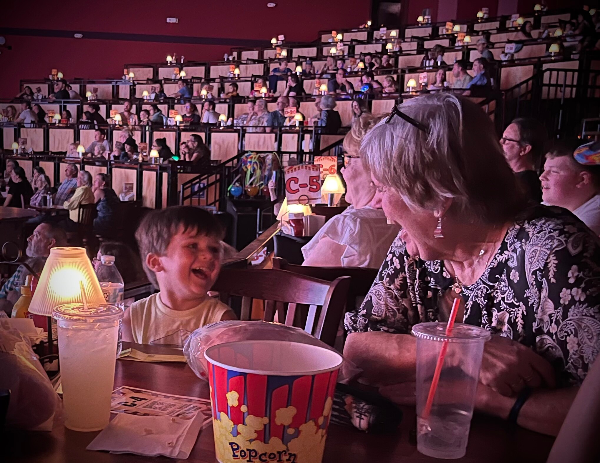 Crowded theater or dinner venue with small lamps on tables and audience seated in illuminated rows; foreground shows a smiling boy and a woman.