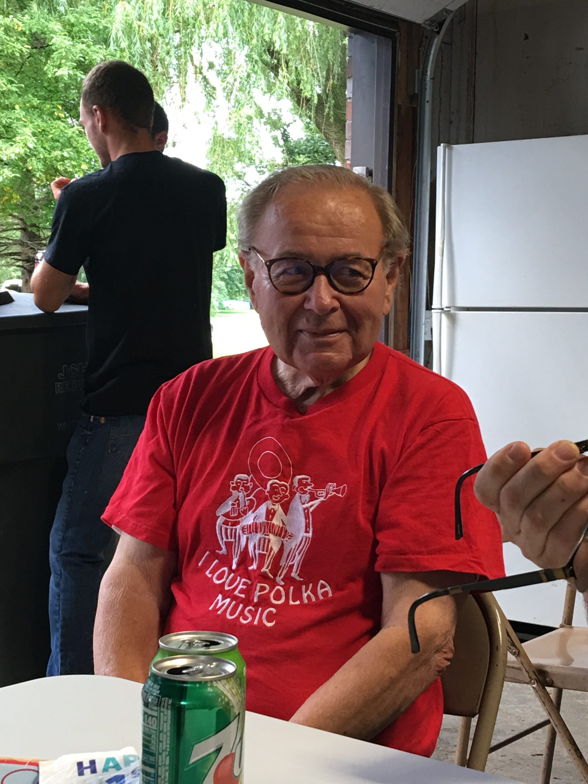 Older man in a red graphic t-shirt sits at a white table with green soda cans, smiling at the camera as another person stands in a garage-like space behind him with greenery outside.