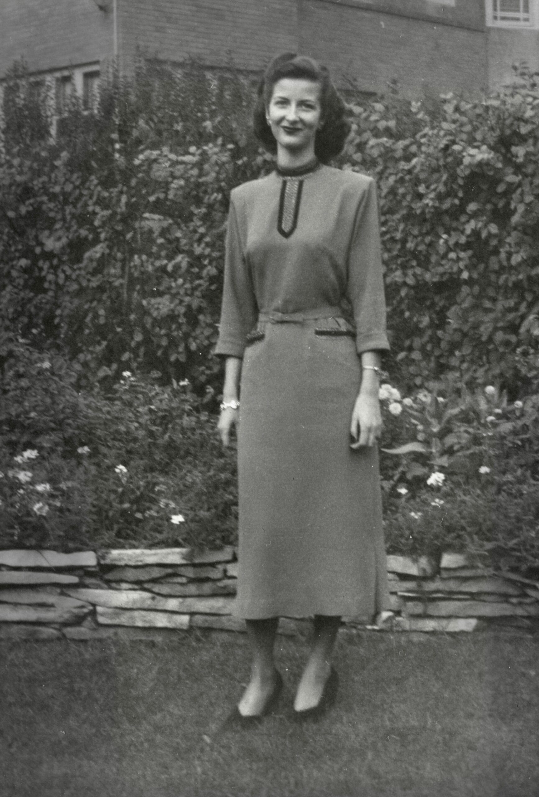 Woman in a vintage mid‑century dress standing in a garden with flowering shrubs and a brick house in the background.