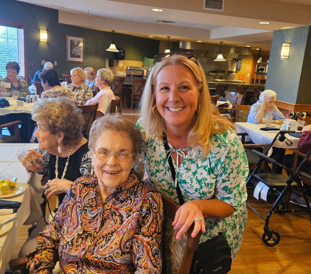 Smiling woman in green patterned blouse standing beside an elderly woman in a colorful blouse at a dining hall table filled with seniors.