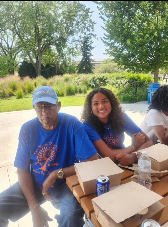 Two people sit at a picnic table outdoors in a park, wearing matching blue shirts, with takeout boxes and beverage cans in front of them on a sunny day.
