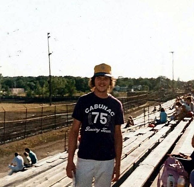 Young man in a tan cap stands on bleachers at a dirt racetrack, wearing a navy shirt with the number 75 and 'Racing Team'.