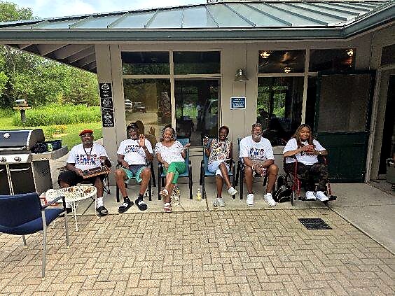 Group of adults seated on chairs outside a storefront, wearing matching white shirts for a community event, on a sunny day.