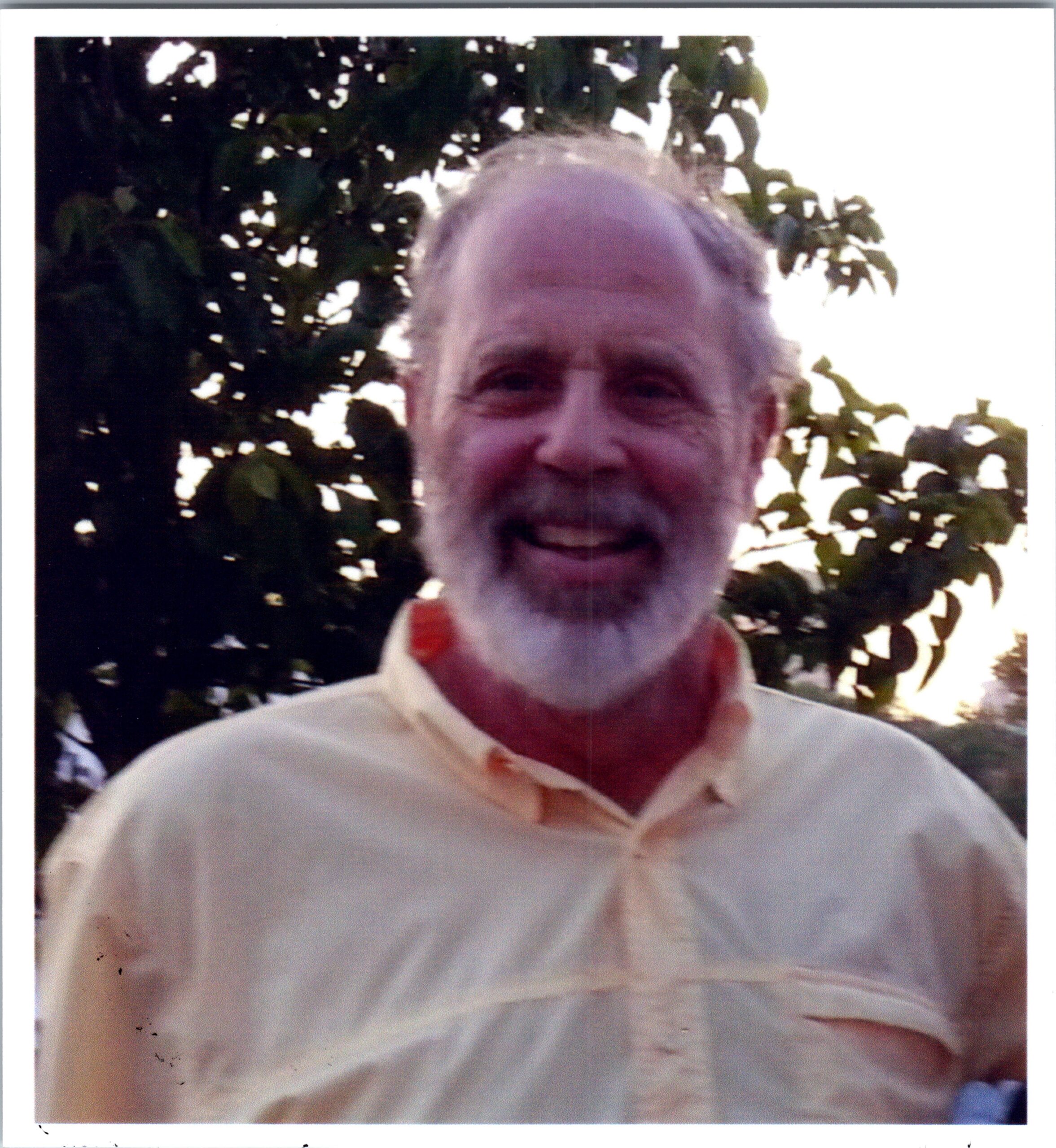 Close-up of a smiling older man with a gray beard outdoors among green trees in a sunny setting.