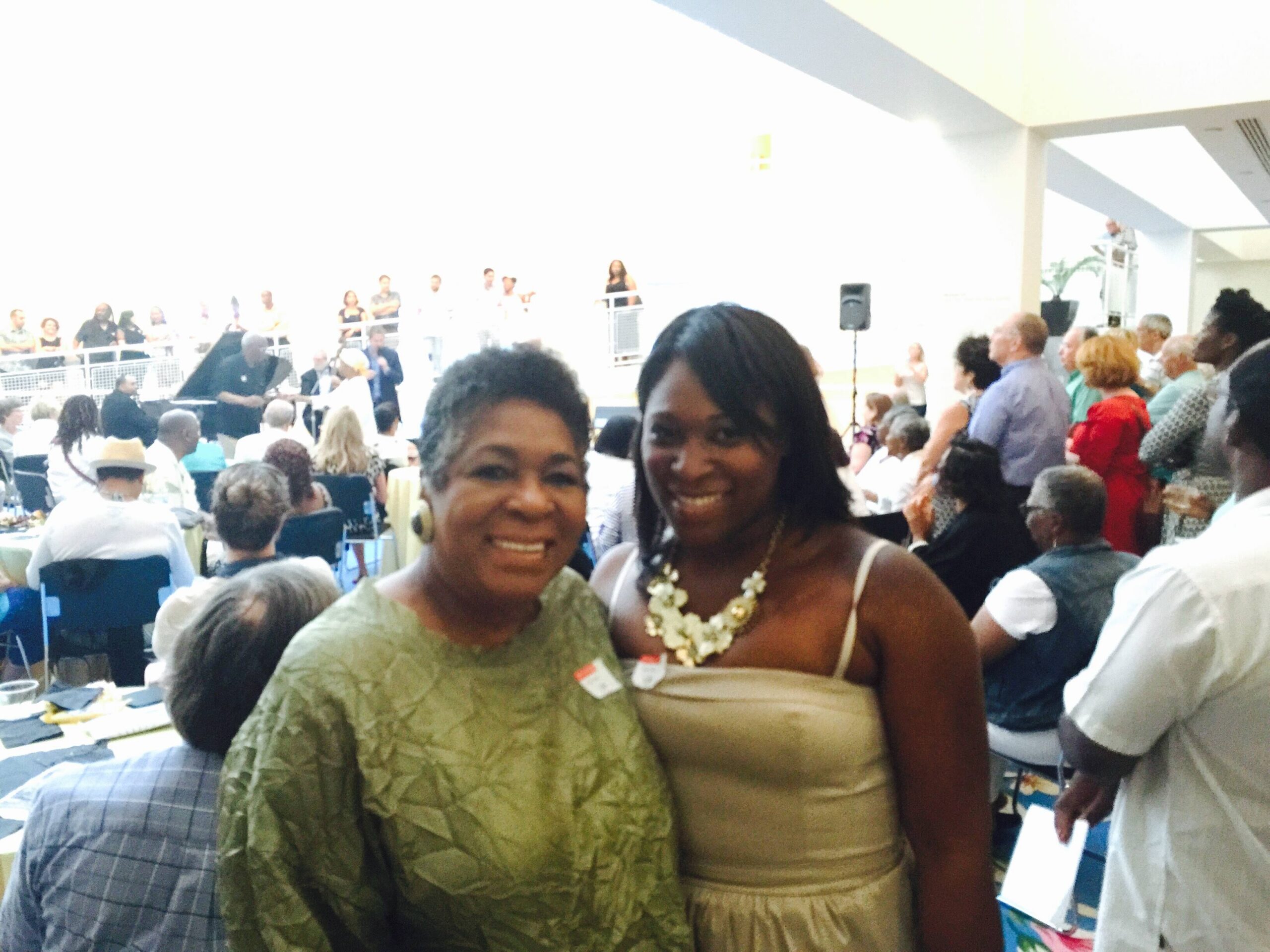 Two smiling women pose for a photo in a crowded indoor event, with guests seated and mingling in the background.