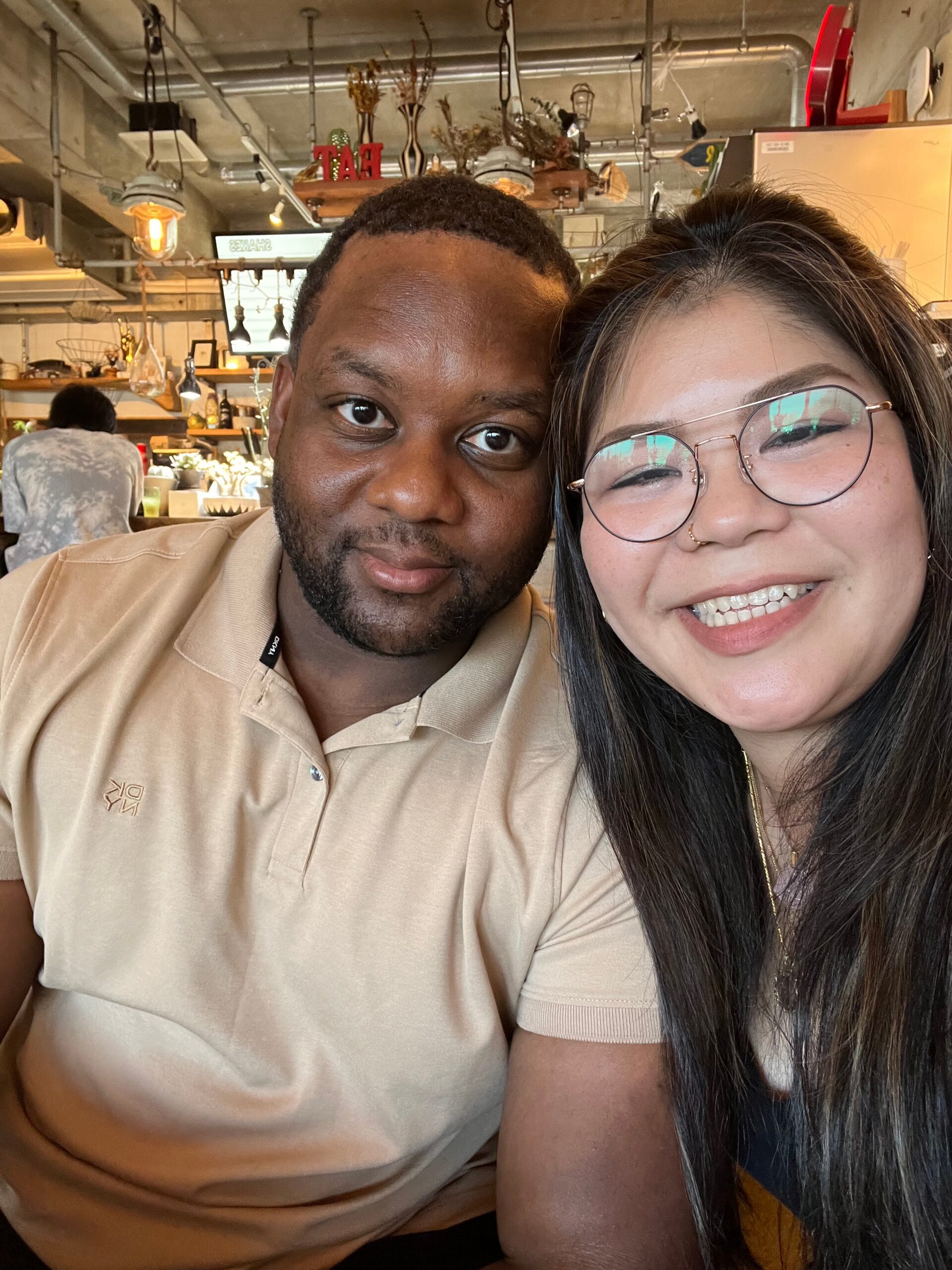 Close-up selfie of a man and a woman smiling in a cozy cafe with exposed pipes and warm lighting in the background. The man wears a beige polo and the woman wears round glasses and braces.