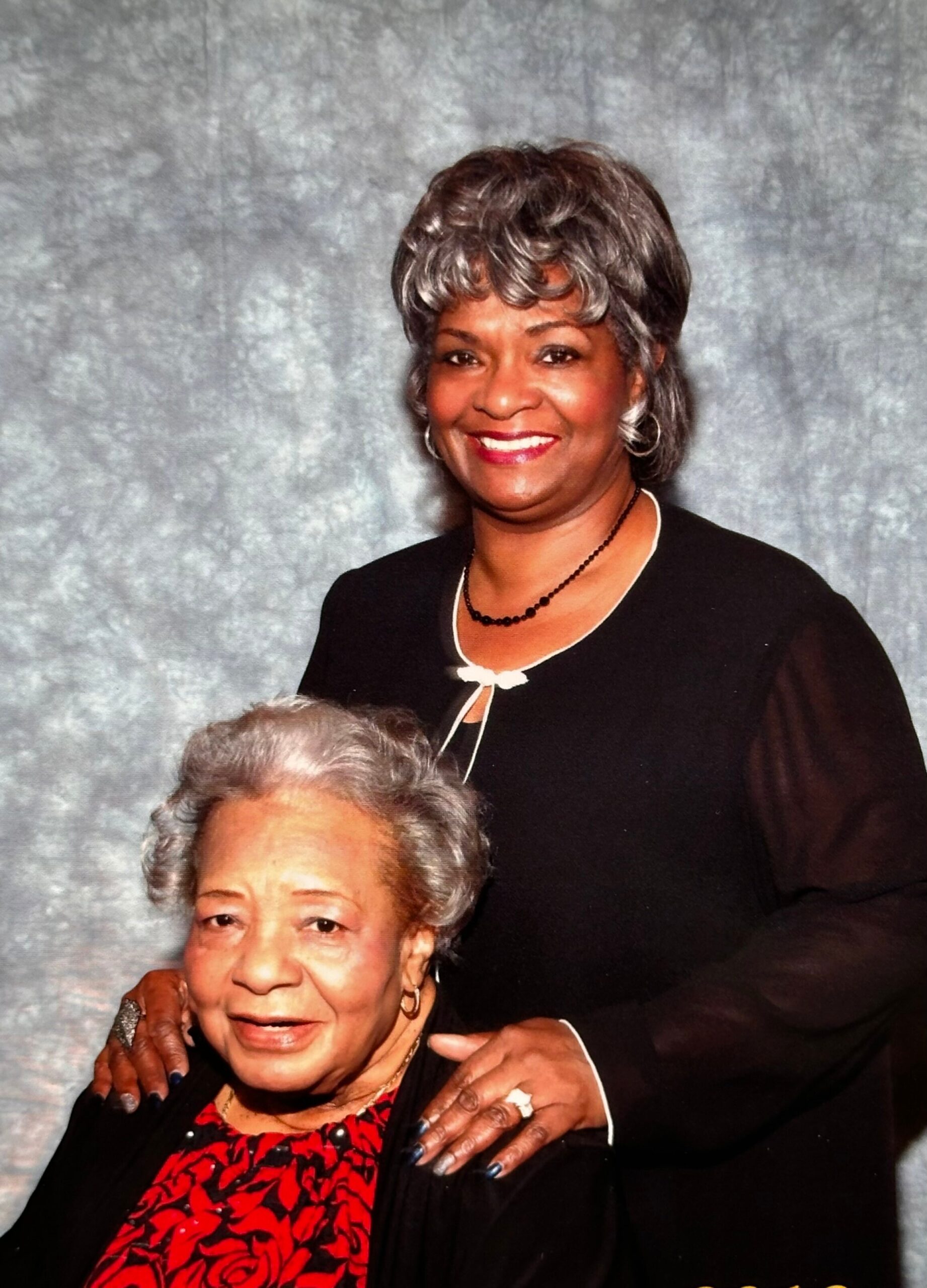 Smiling woman with gray hair standing behind an older woman, both in dark clothing, against a gray backdrop.