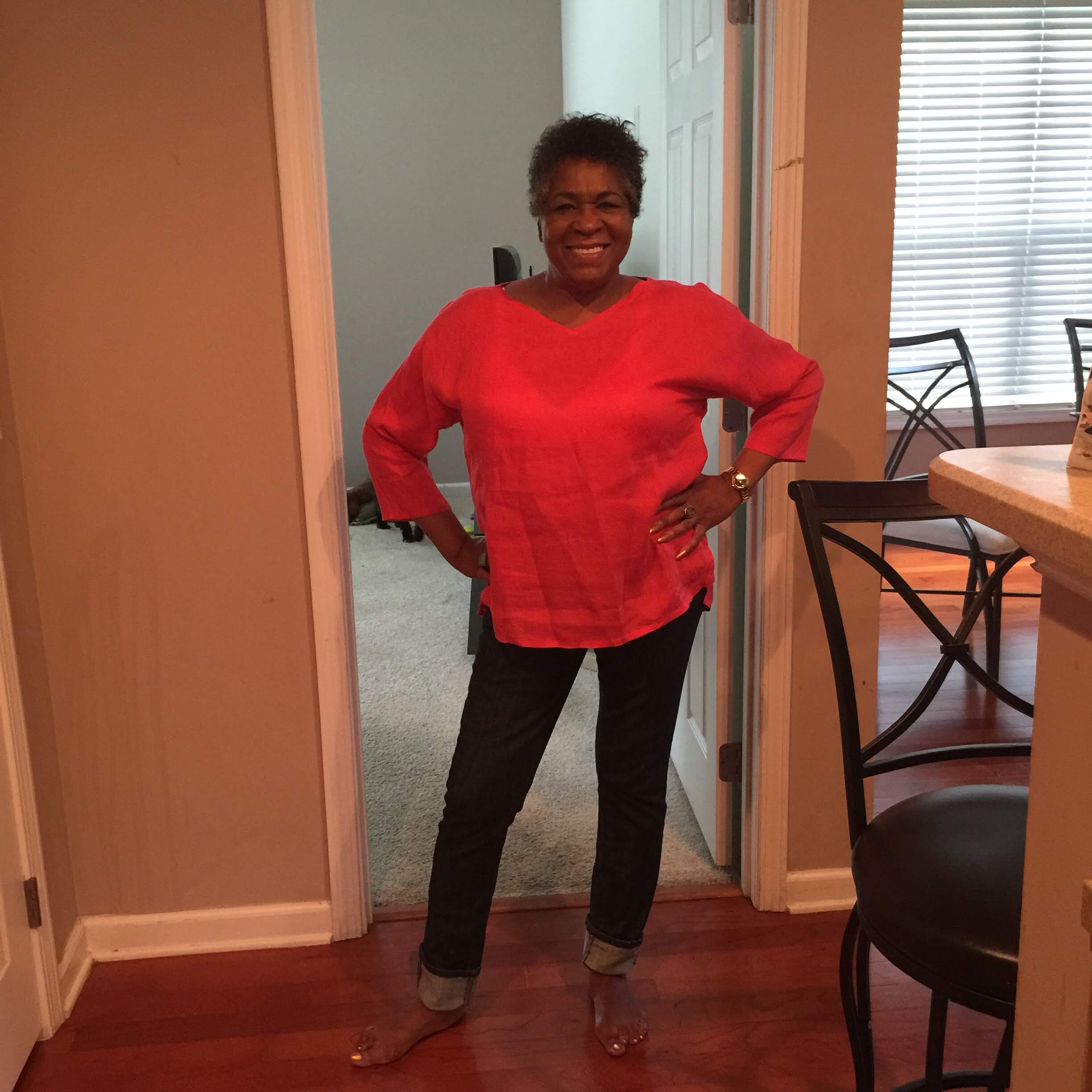 Smiling woman standing in a doorway, wearing a bright red blouse and dark jeans.”