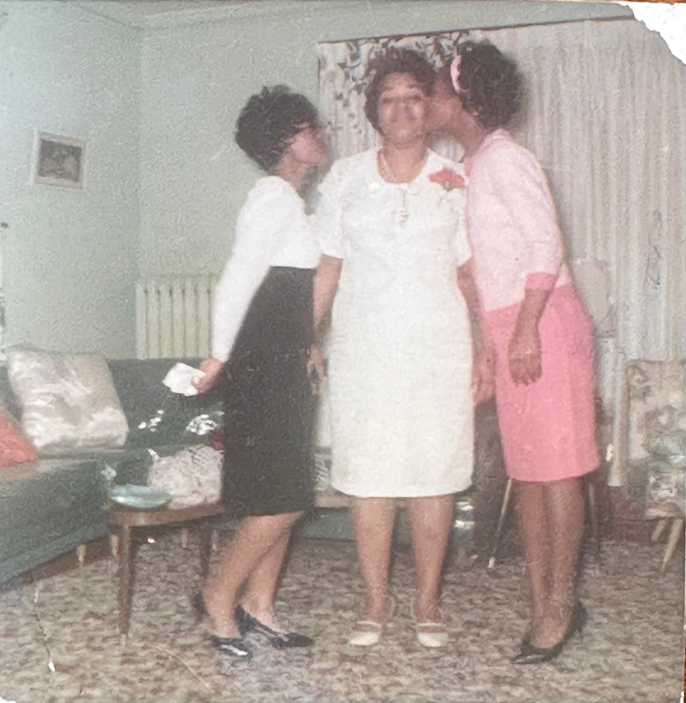Three people indoors; two women kiss a man on the cheek in a casual group photo, living room backdrop with a curtain.