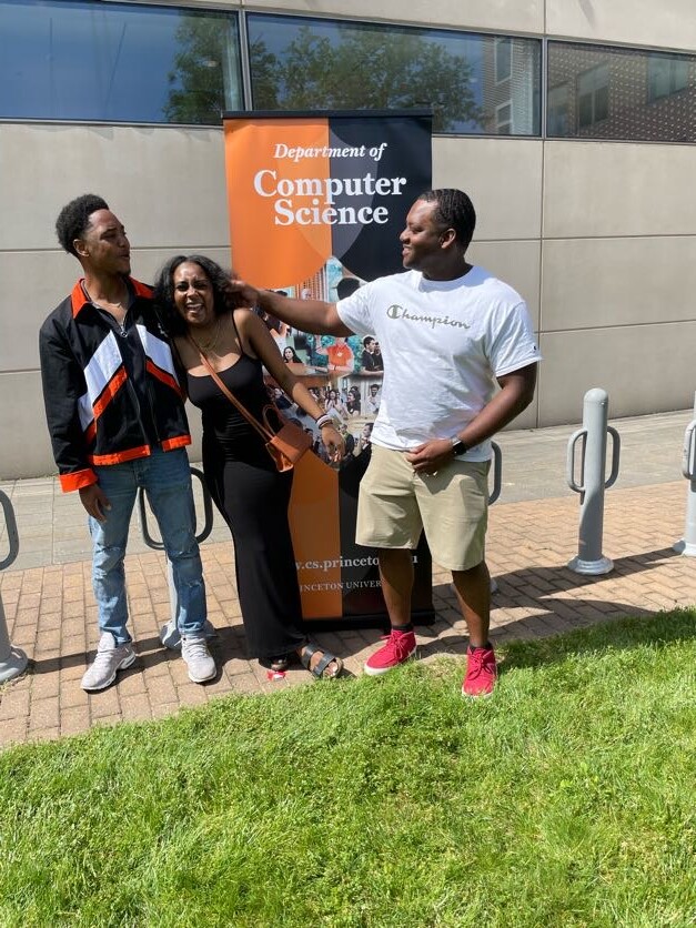 Three people posing outdoors in front of a Department of Computer Science banner; a woman between two men smiling and interacting.