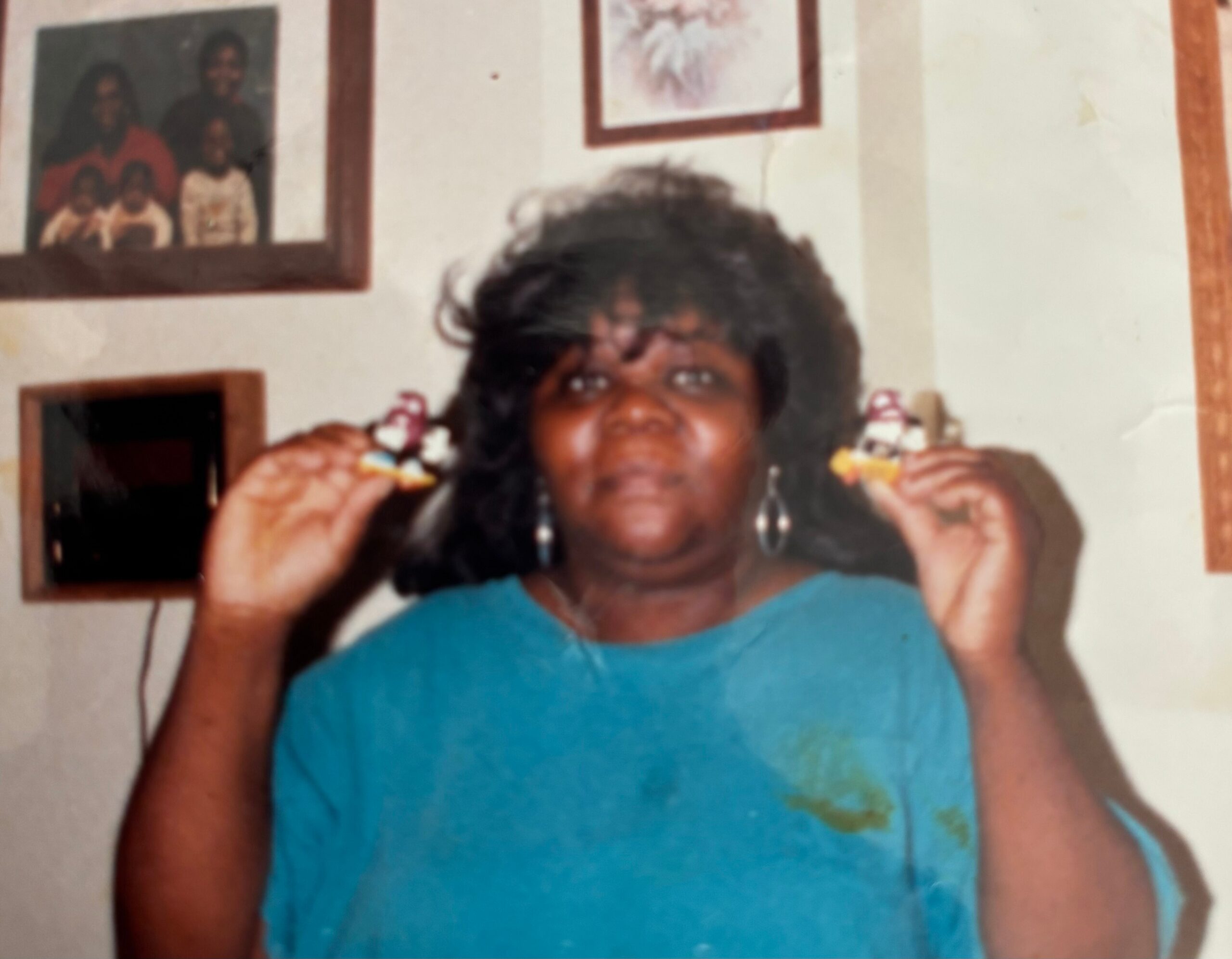 Person with big, dark hair wearing a teal shirt, indoors, holding a small snack near their mouth with framed pictures on the wall behind.