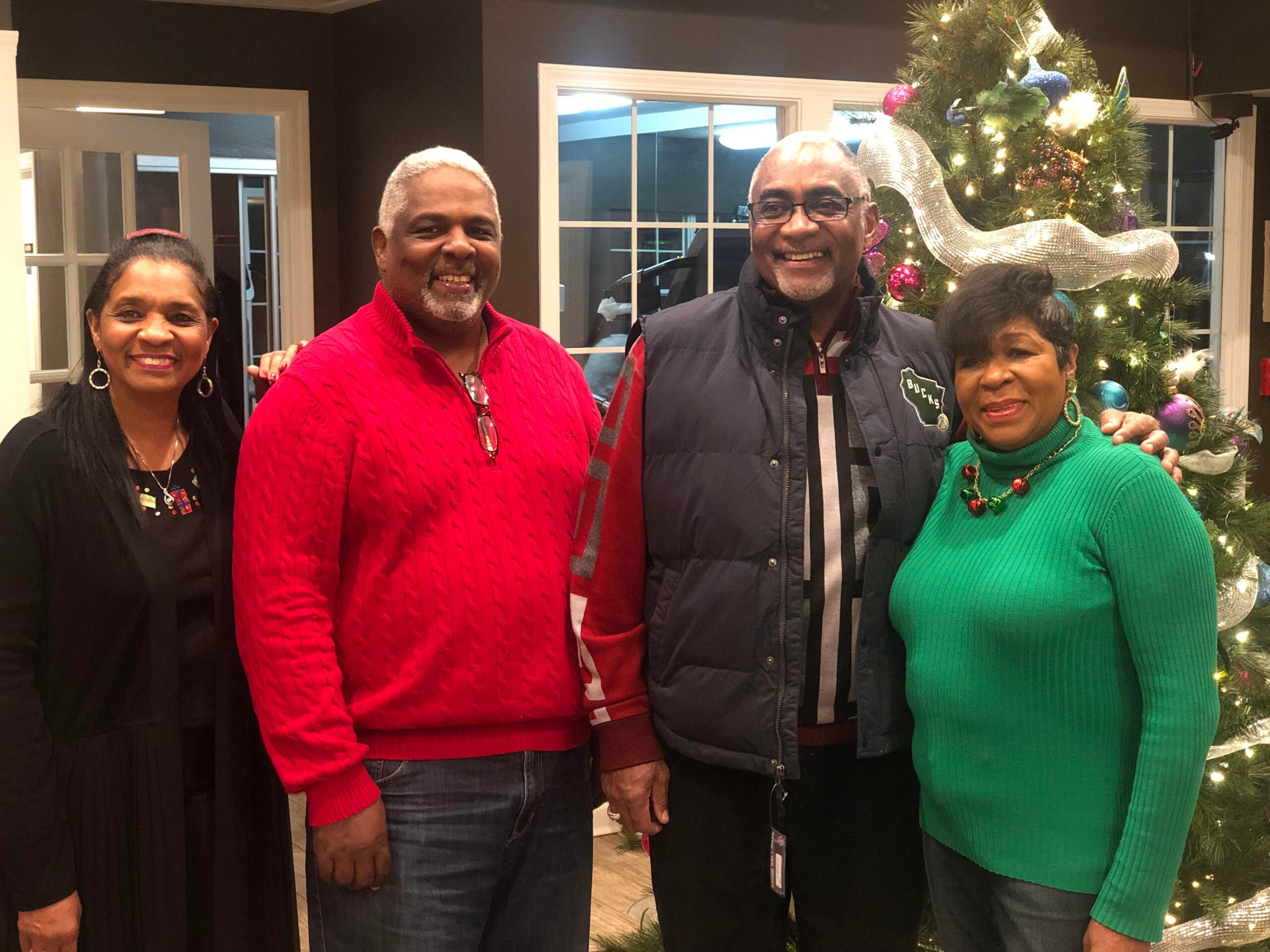 Four adults pose together indoors next to a decorated Christmas tree with lights and ornaments.