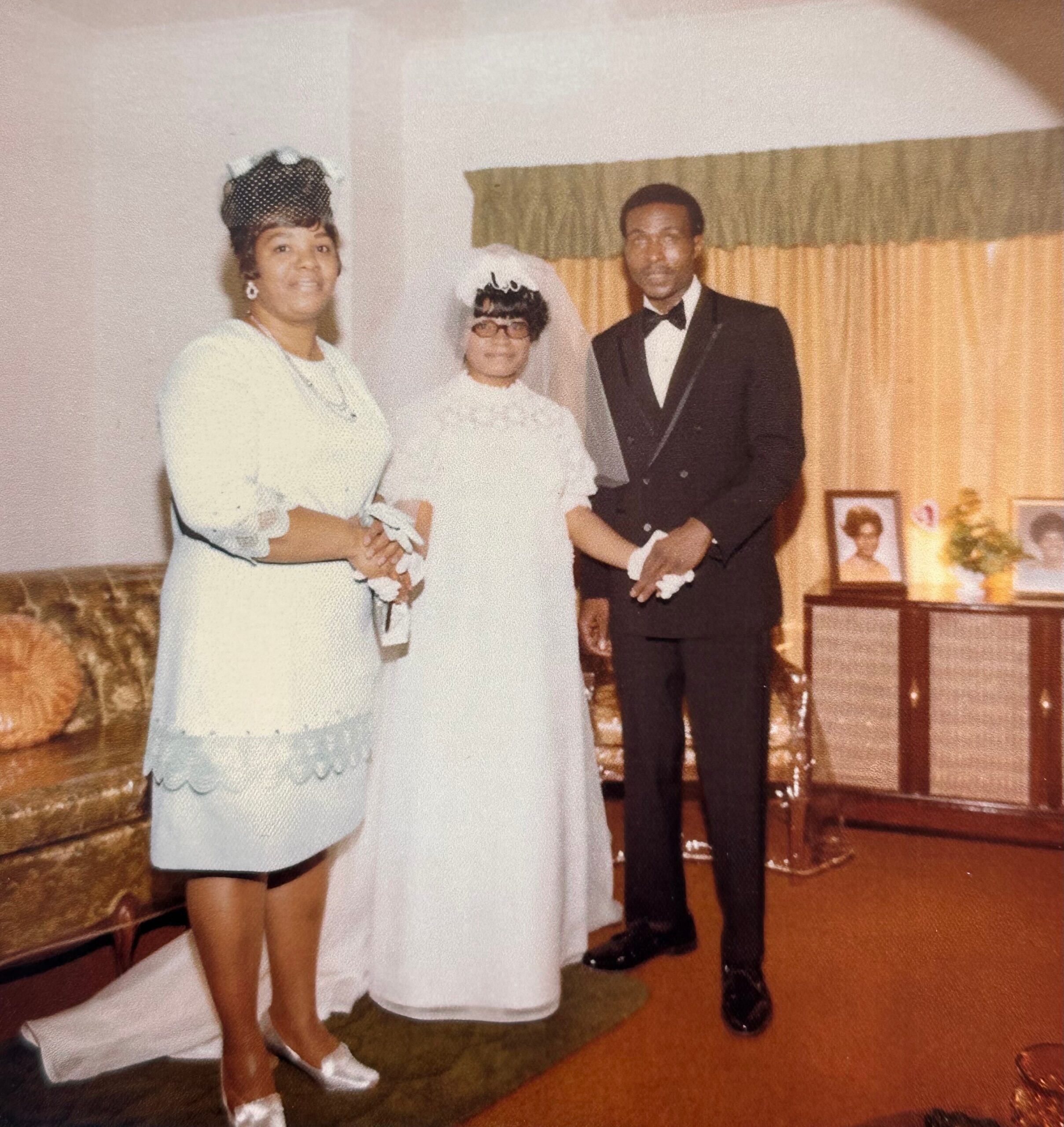 Bride in a white wedding dress and veil stands with a smiling woman in pale dress and a man in a tuxedo indoors.