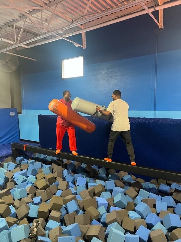 Two men in a gym balance on a narrow beam over a foam pit, exchanging large foam batons.