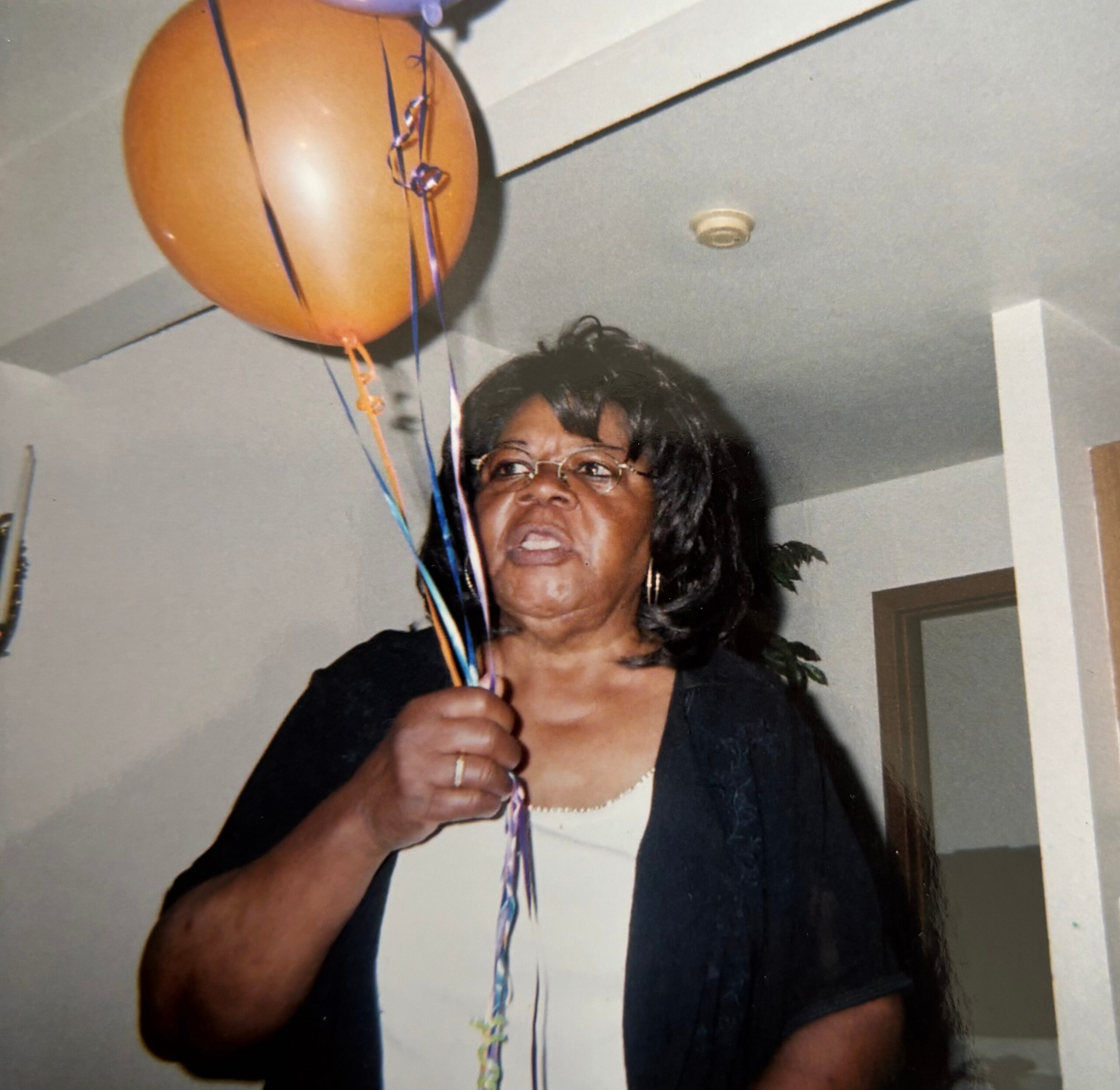 Older woman with short dark hair and glasses holding colorful balloons indoors, smiling slightly.