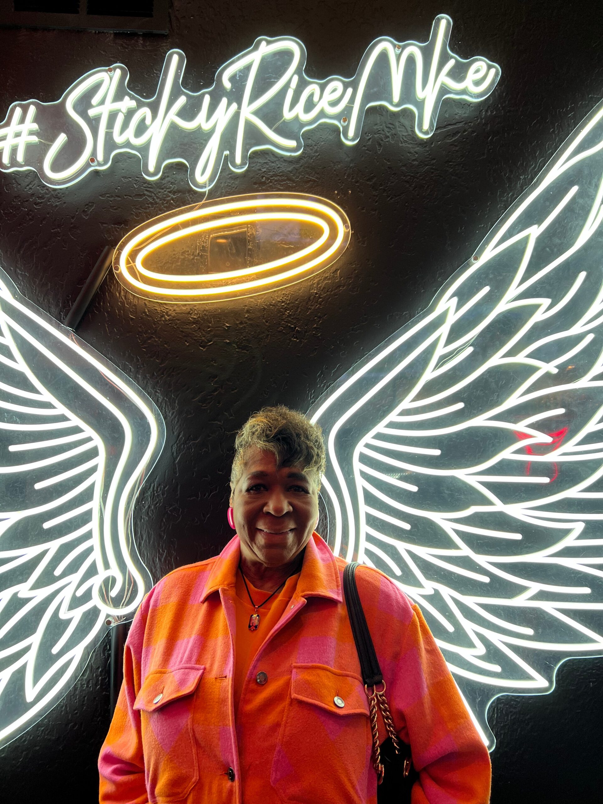 Person smiling in an orange-pink plaid shirt stands in front of neon wing mural with a glowing halo above, on a dark wall background.