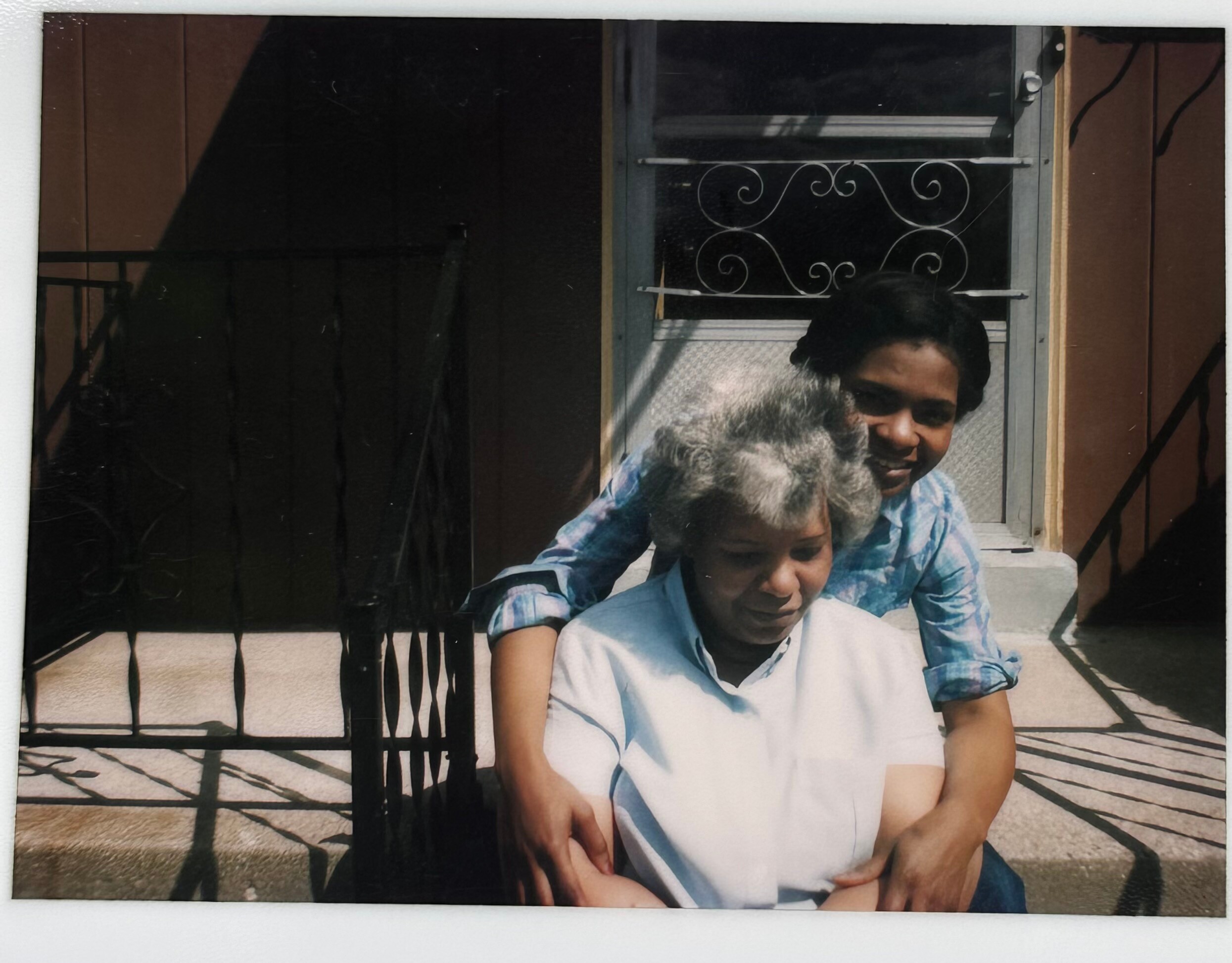 Two people smile on a porch: a man in a blue shirt with a child on his lap in front of a window with a decorative iron grille.