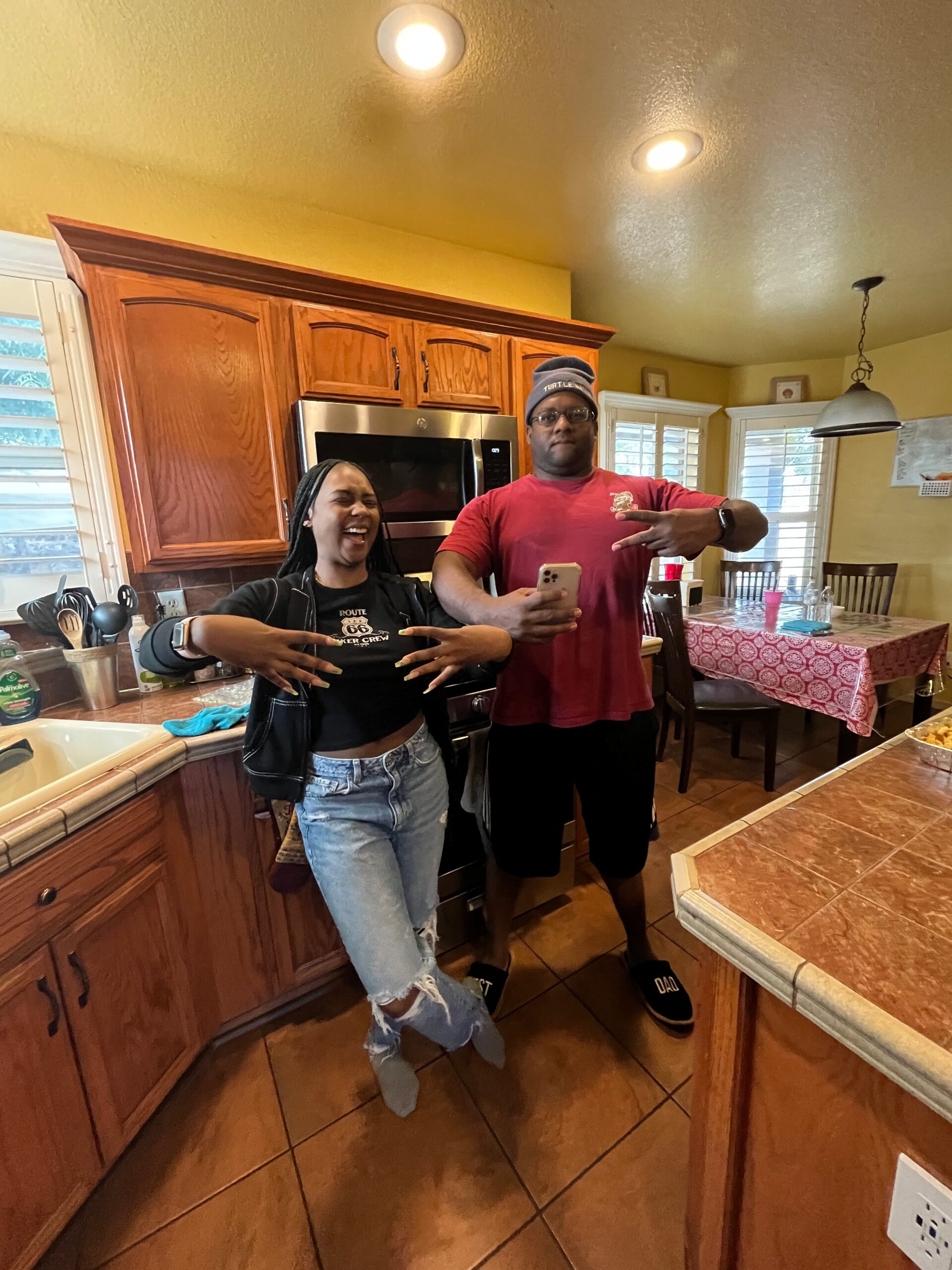 Two friends posing playfully in a kitchen, showing peace signs and smiles near the stove, with wooden cabinets and a dining table in the background.