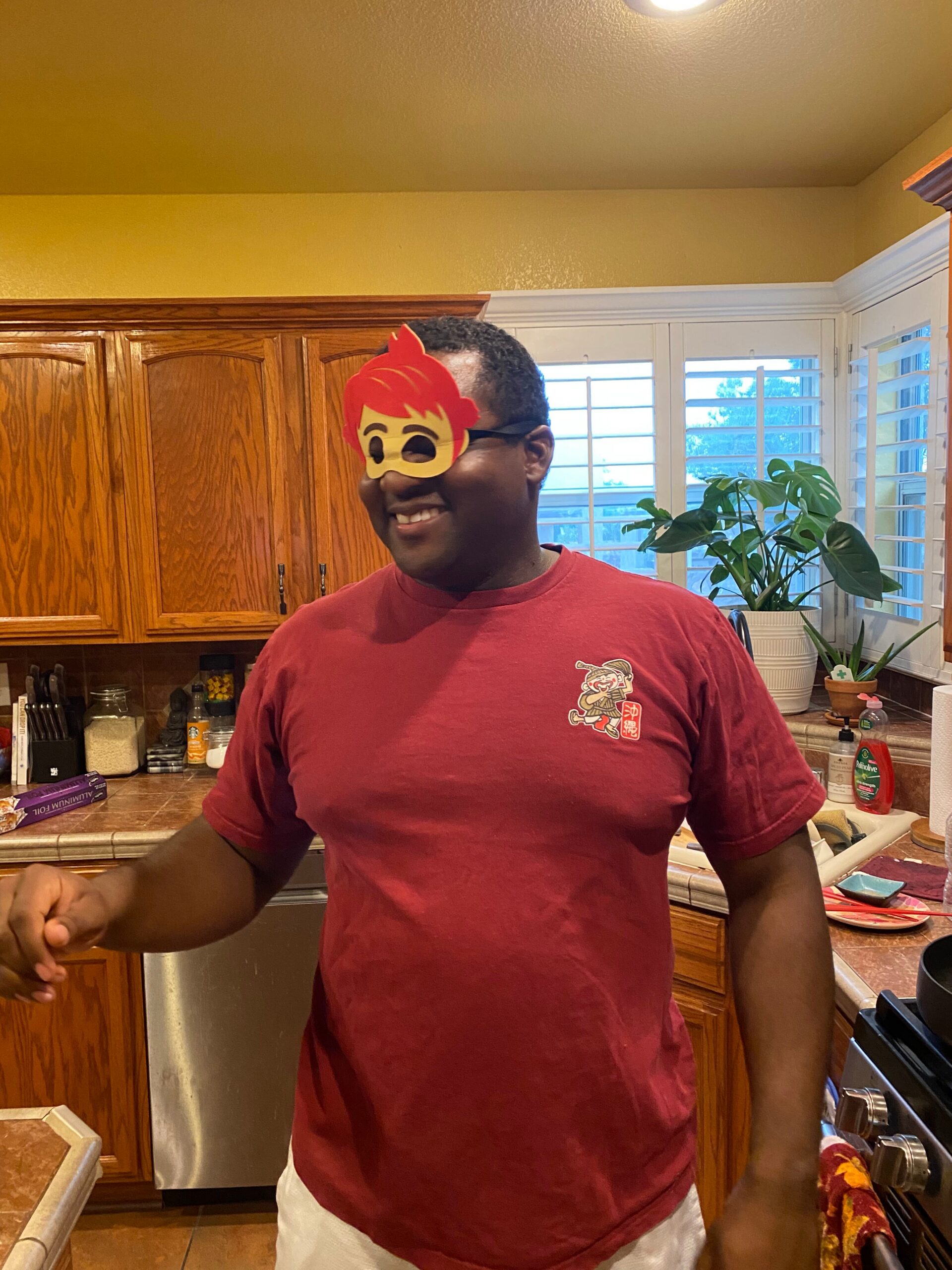 Man smiling in a red shirt wearing a red mask over his eyes, standing in a kitchen with wooden cabinets and a plant in the background