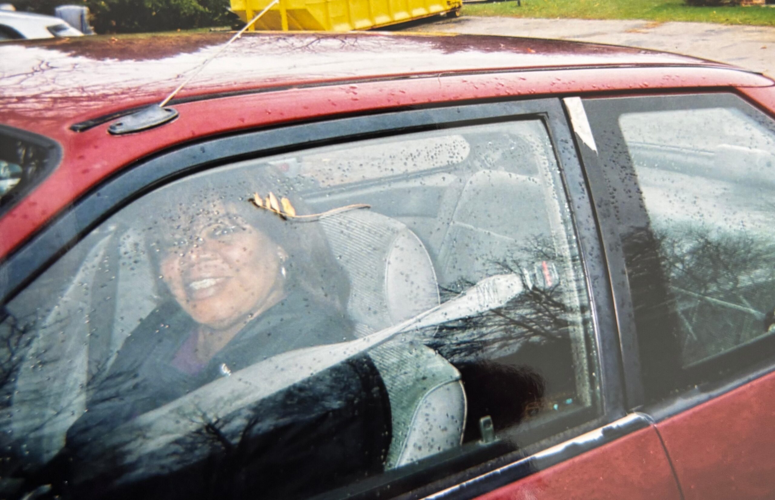 Smiling person in a red car, seen through a rain-speckled windshield and side window, outdoors.