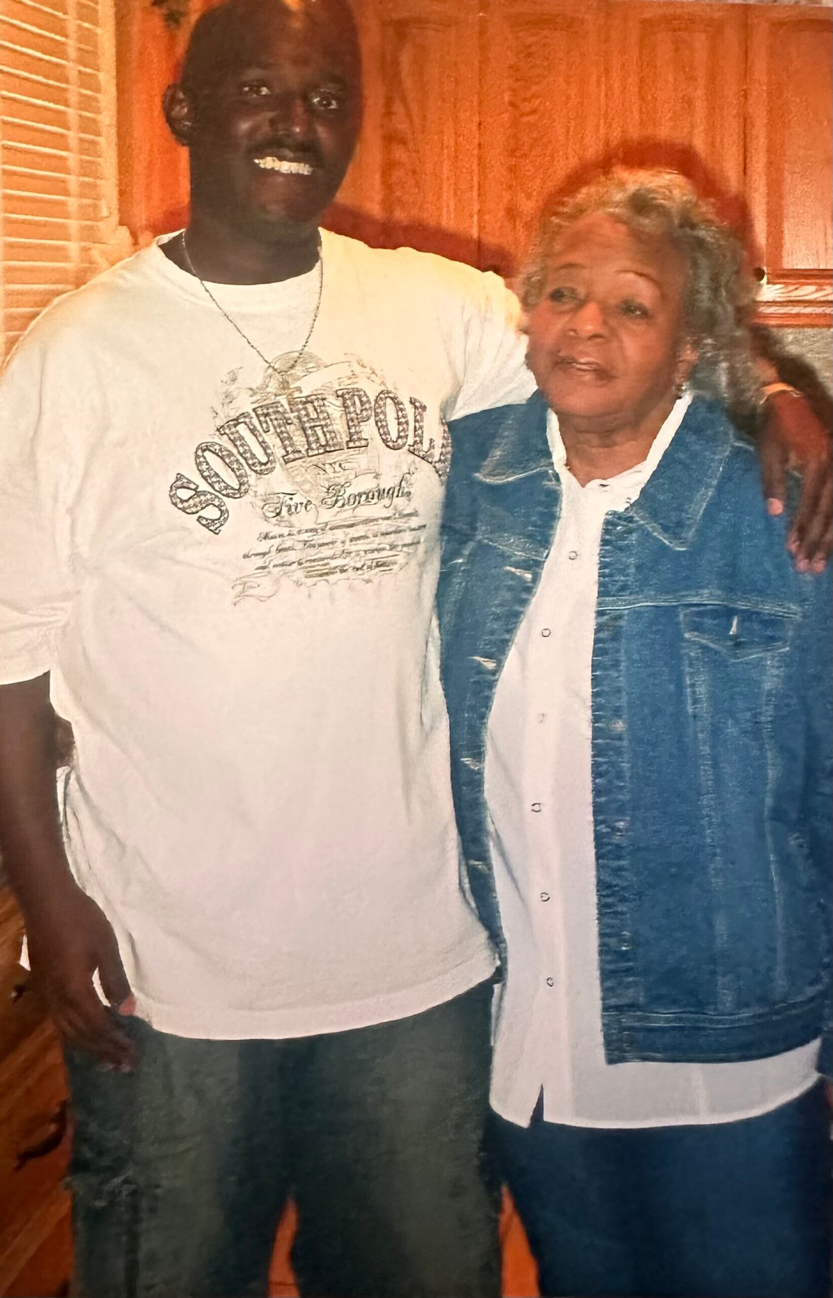 Smiling man in a white Southpole t-shirt with a necklace stands next to an elderly woman in a denim jacket, arm around her, posing indoors.