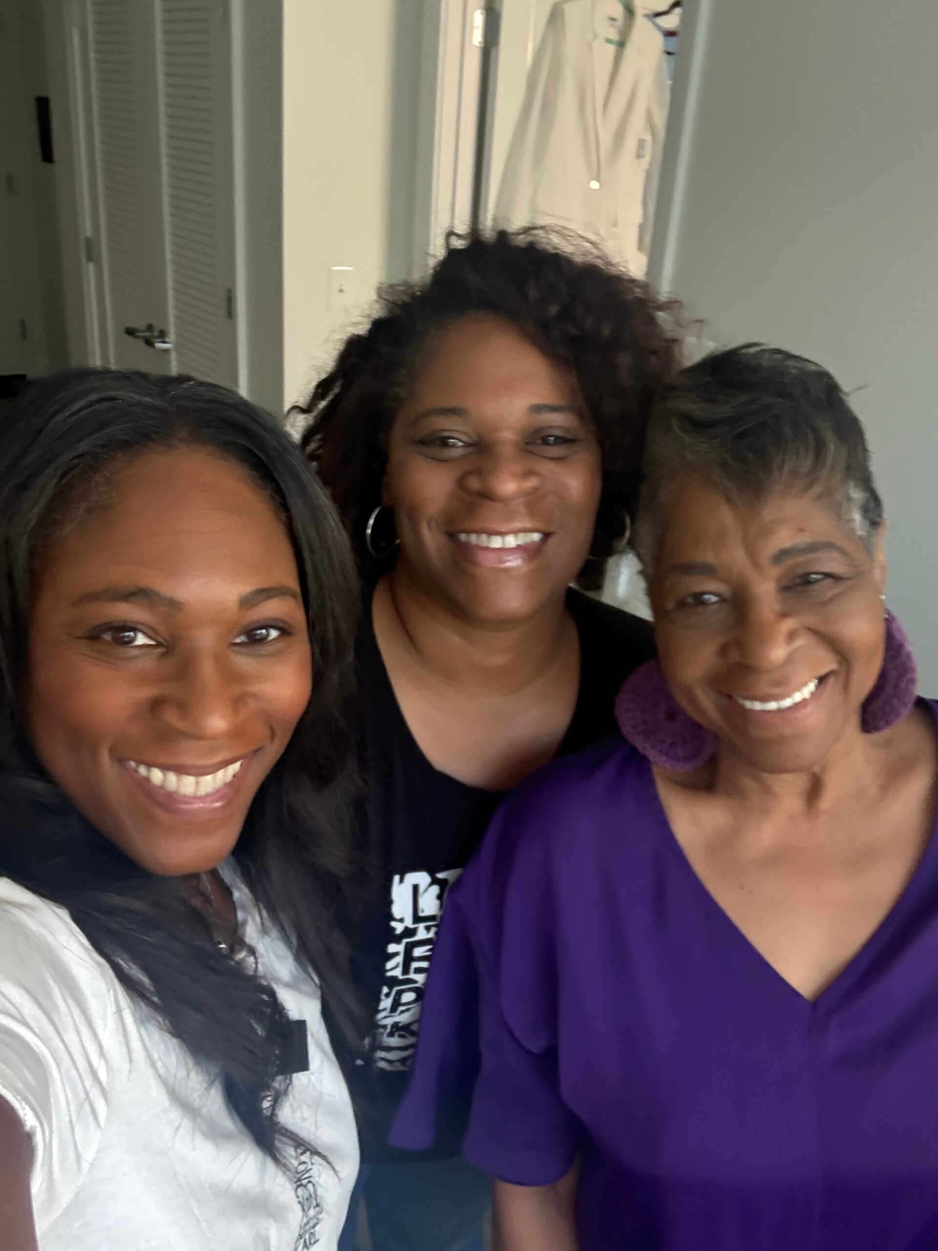 Three women smiling for a selfie in a hallway, two in the back and one in the front wearing a white top, others in darker clothing.