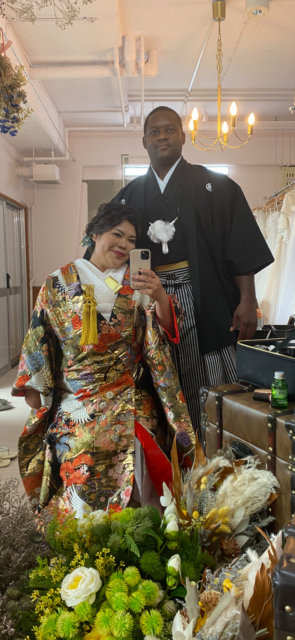 Couple in a dressing room posing for a selfie; woman wears a colorful, ornate kimono, man in black kimono with striped hakama; flowers in foreground.