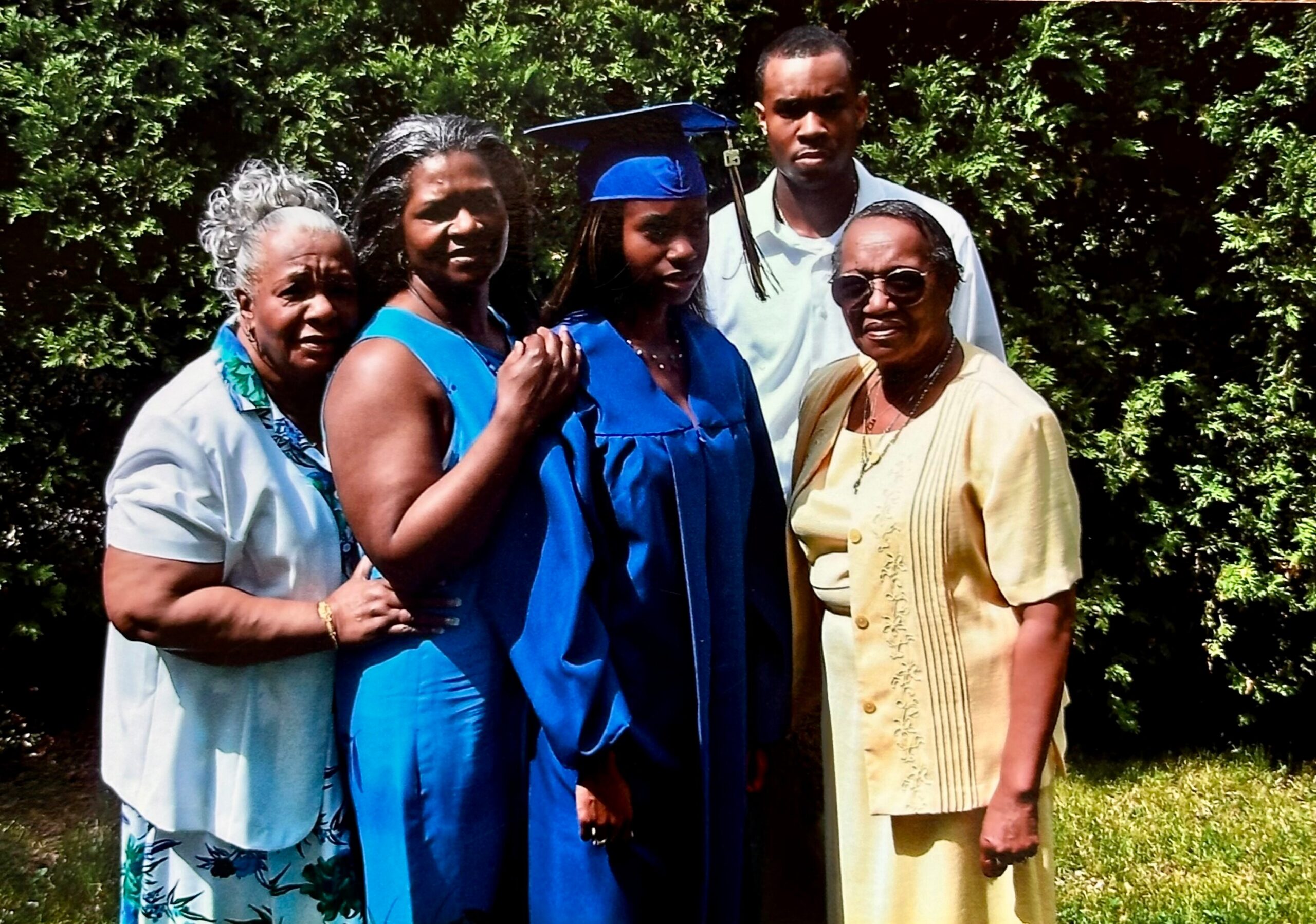 Group of five people posing outdoors for a graduation photo; a young woman in a blue cap and gown stands center, flanked by family members in light-colored outfits against a green hedge backdrop.