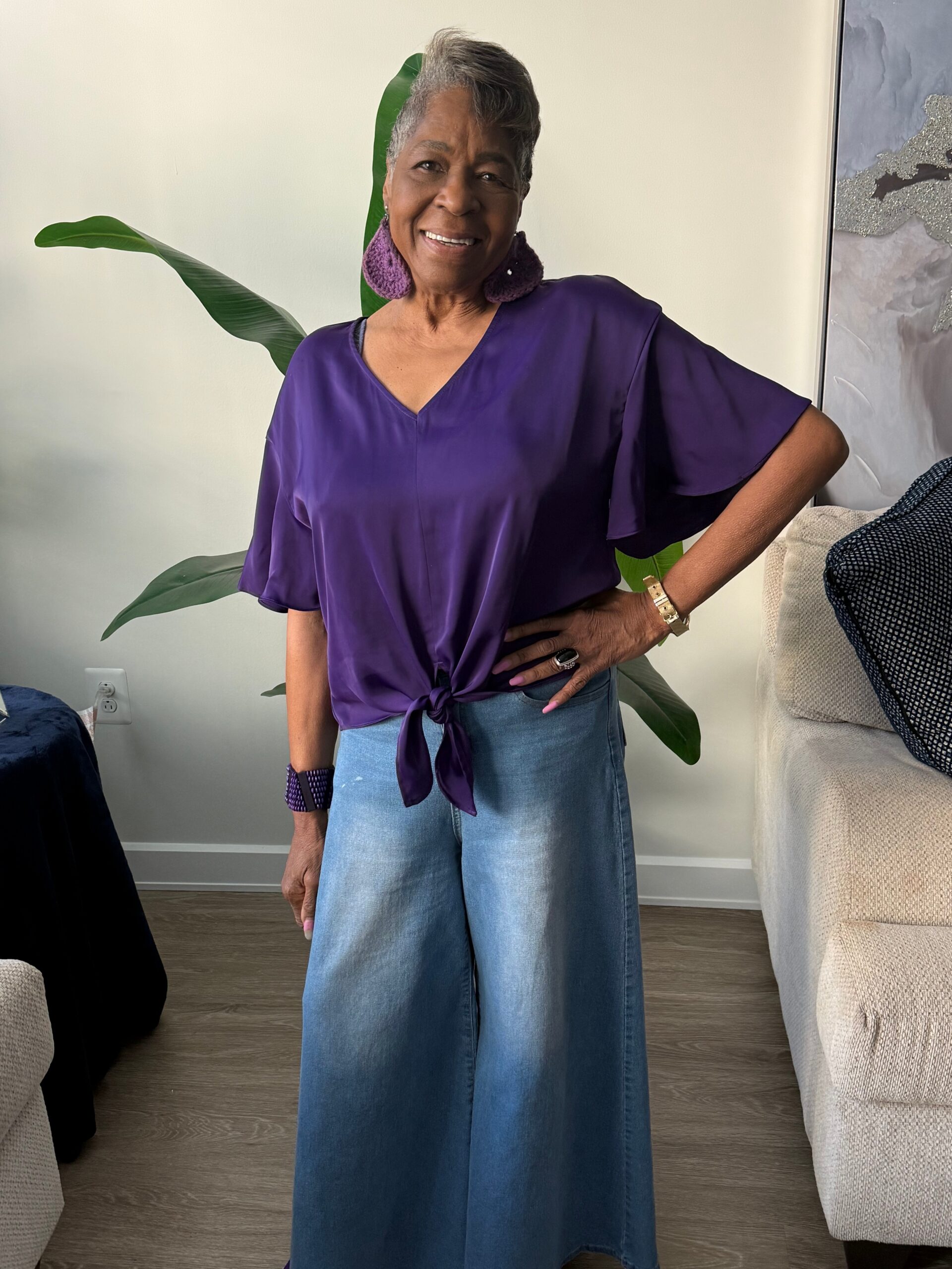 Smiling woman with short gray hair wearing a purple top tied at the waist, wide-leg jeans, purple earrings, and gold watch, standing with one hand on her hip in a living room with plants nearby.
