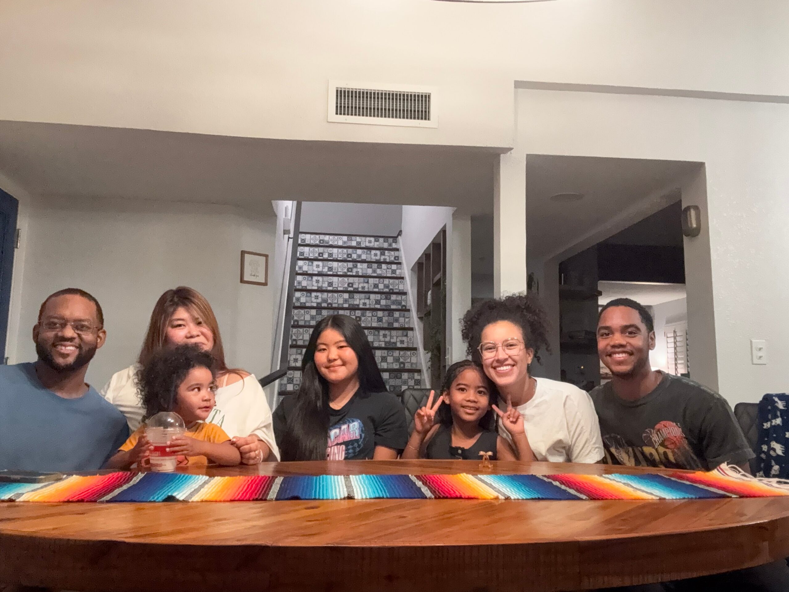 Group of eight people smiling at the camera around a long wooden table with a rainbow-striped table runner indoors.