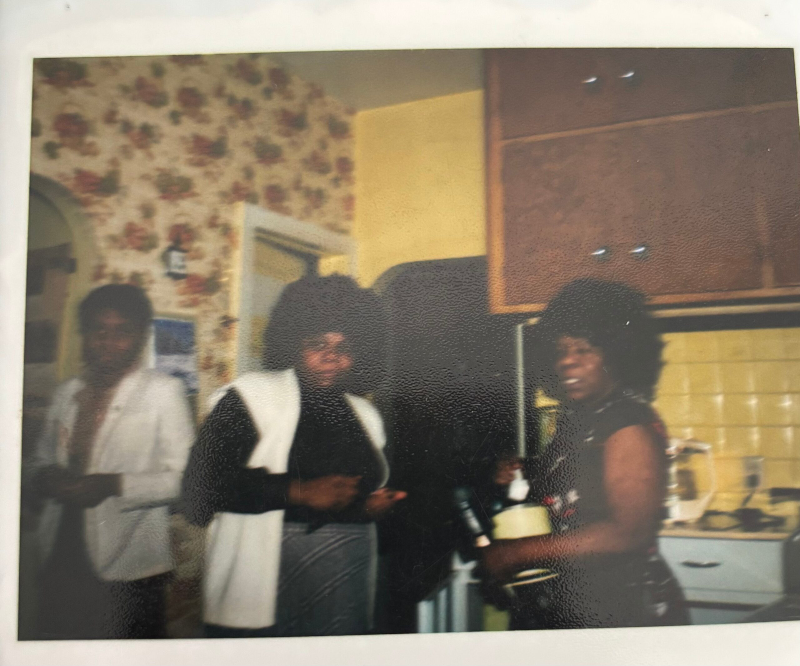 Group of people standing indoors in a room with floral wallpaper on the left and wooden cabinets on the right; photo is blurry and low-resolution.