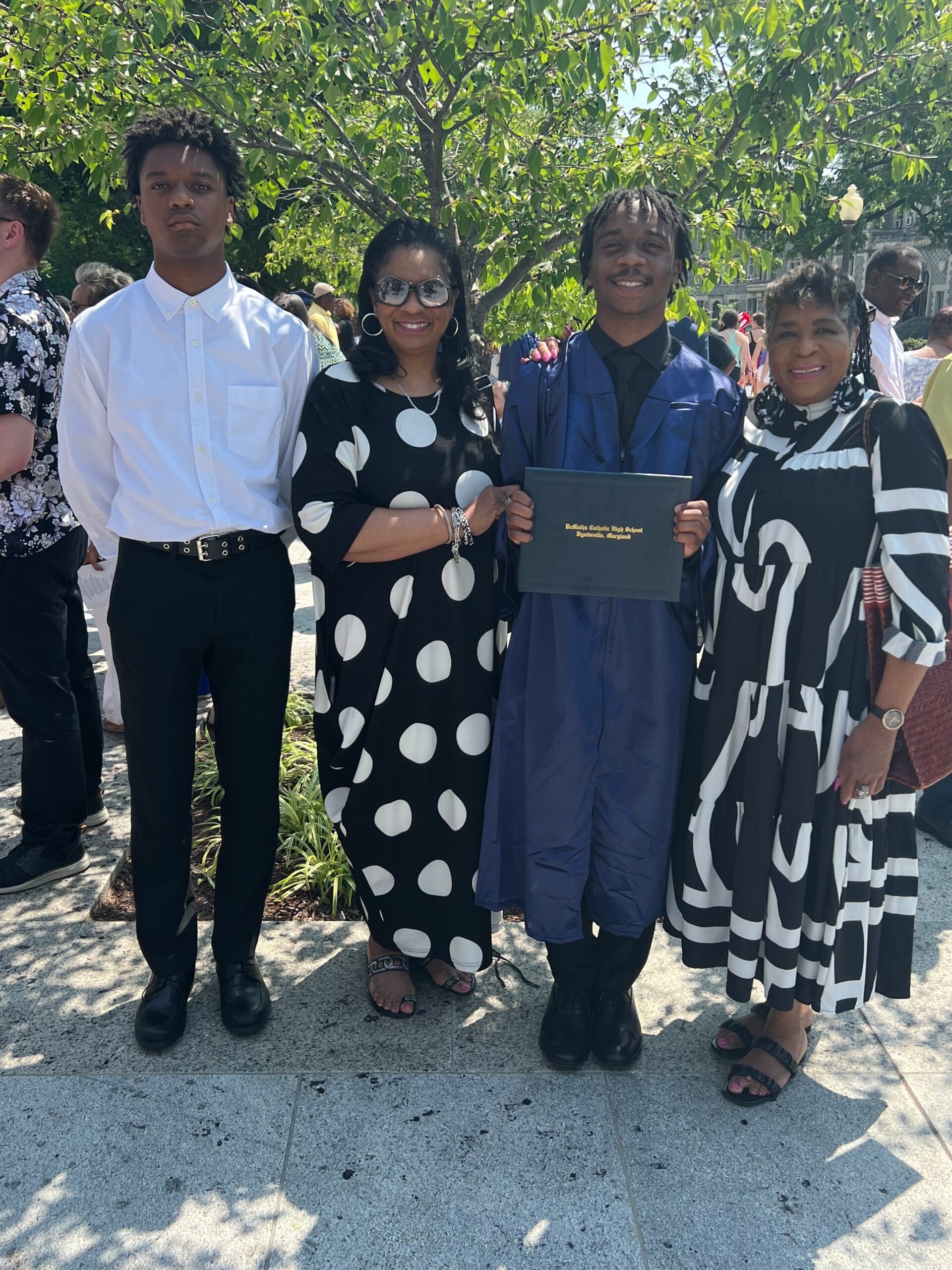 Graduate in a blue cap and gown holds a diploma while posing with family outdoors under a tree.