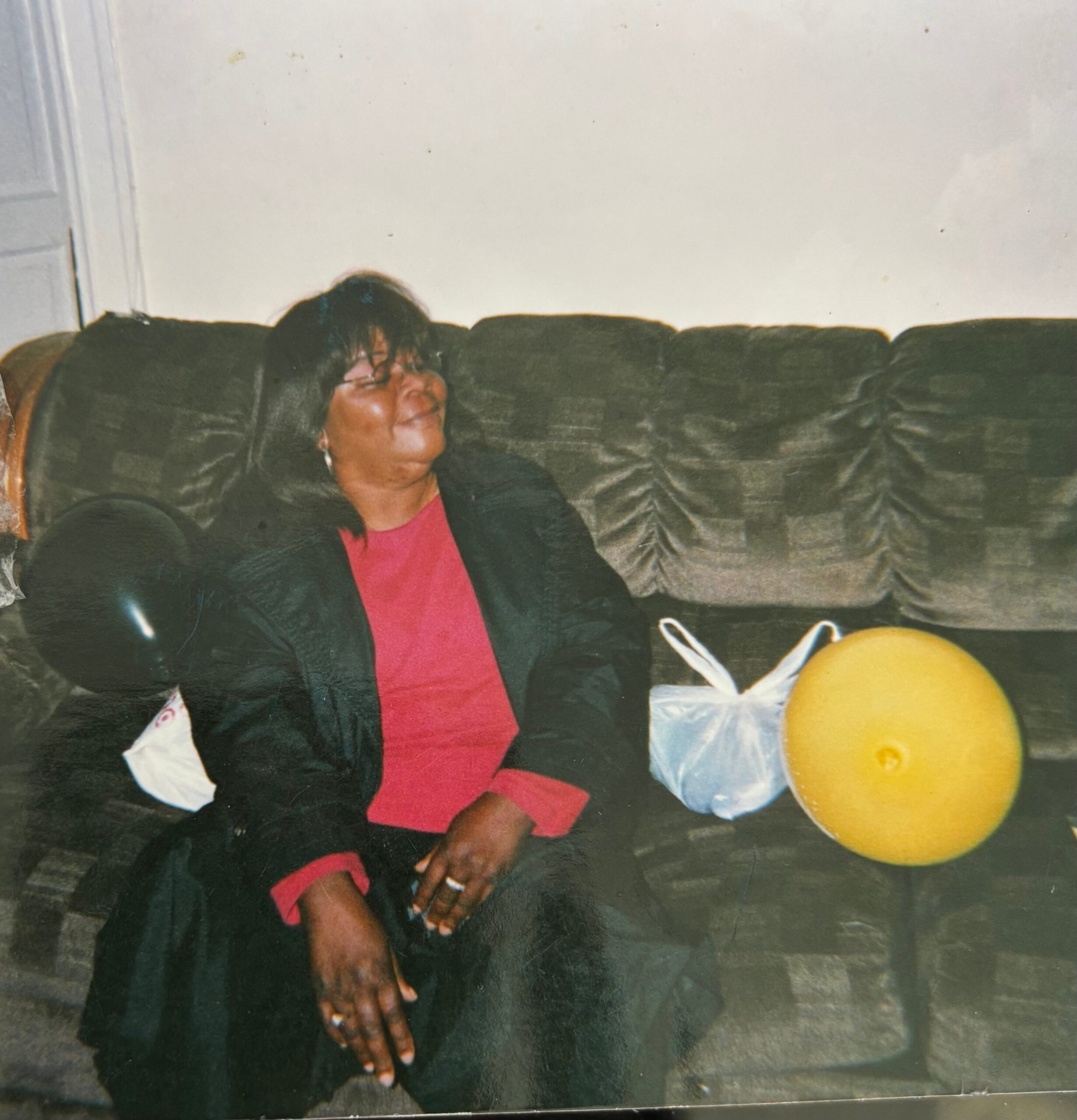 Person seated on a dark couch indoors, wearing a red shirt and black blazer, smiling with eyes closed; a yellow ball sits on the floor nearby.