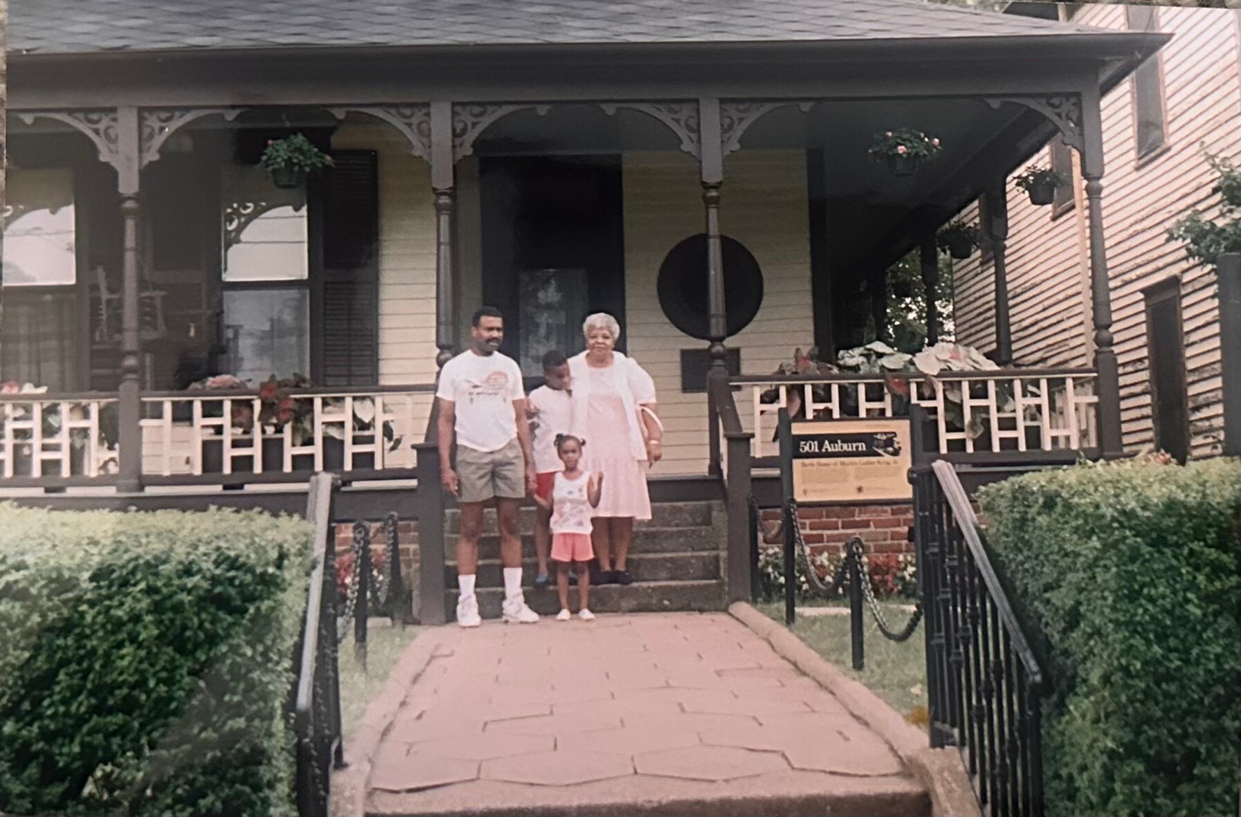 Family of four posing on the front steps of a cream-colored house with a decorative dark porch, address sign 501 Auburn visible on the railing.