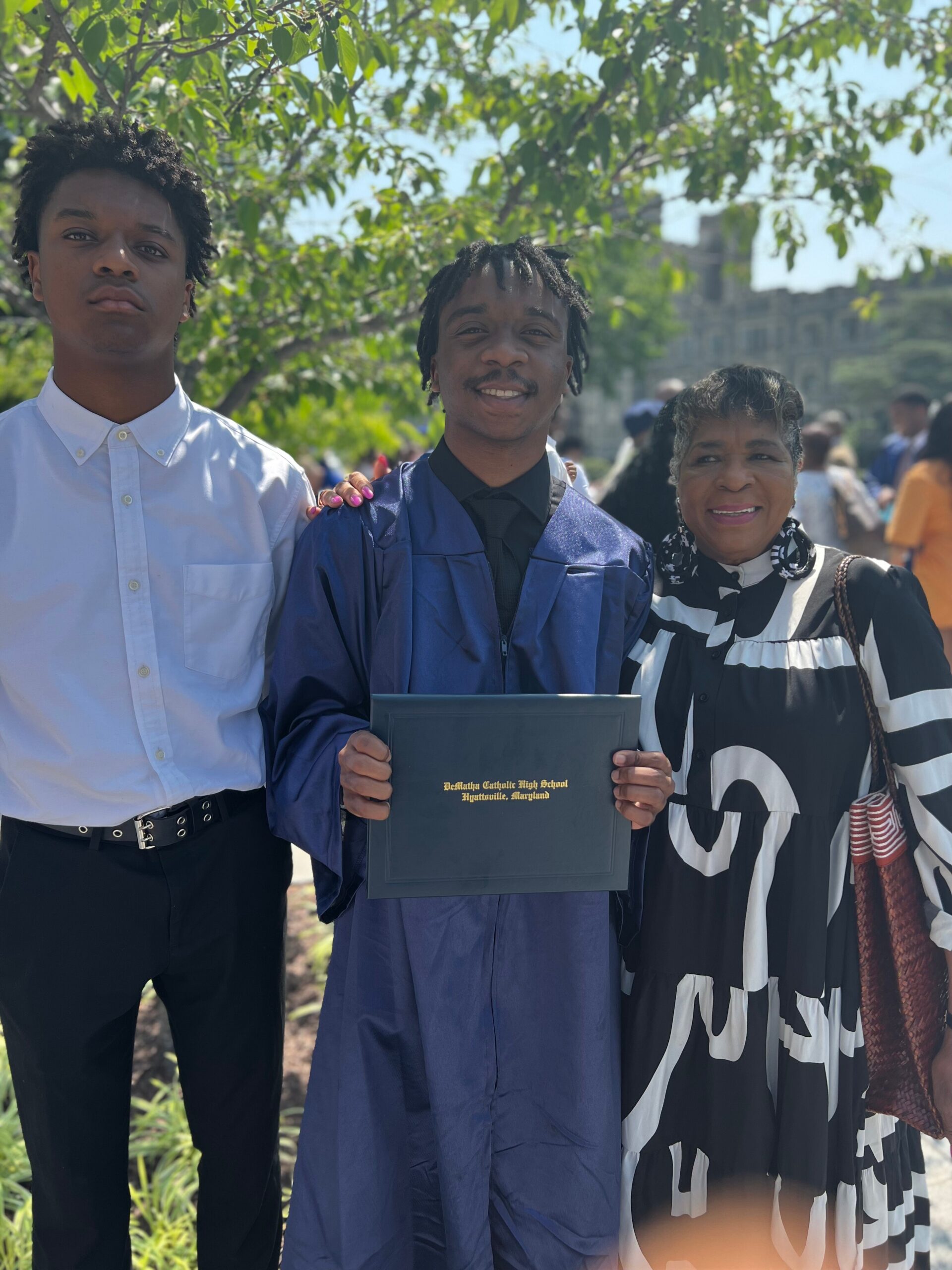Graduate in blue gown holding a diploma, flanked by a young man in a white shirt and a smiling woman outdoors under trees.
