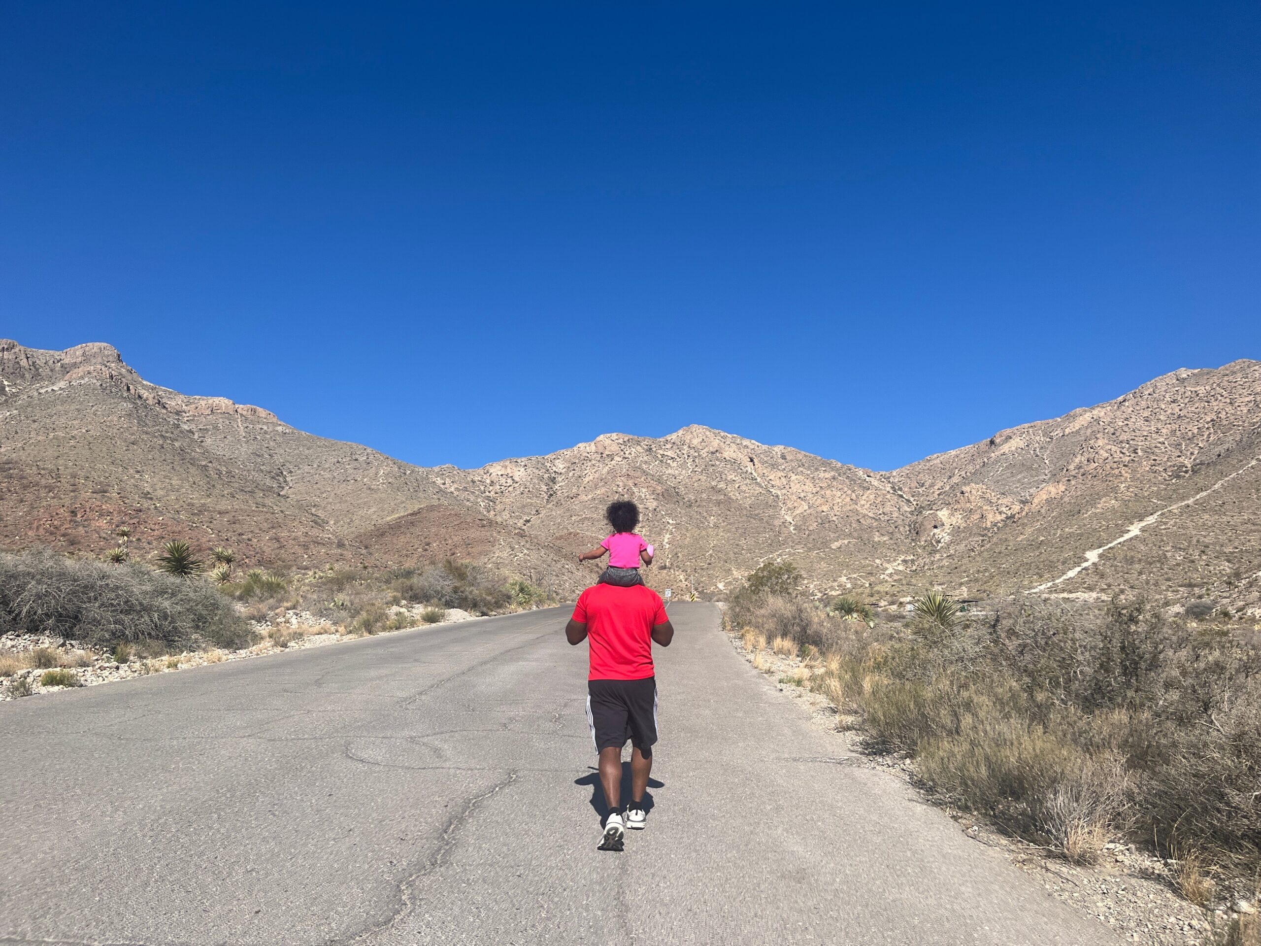 Back view of a person in a red shirt running on a desert road with a child in a pink shirt on their shoulders; rocky hills and a clear blue sky in the background.