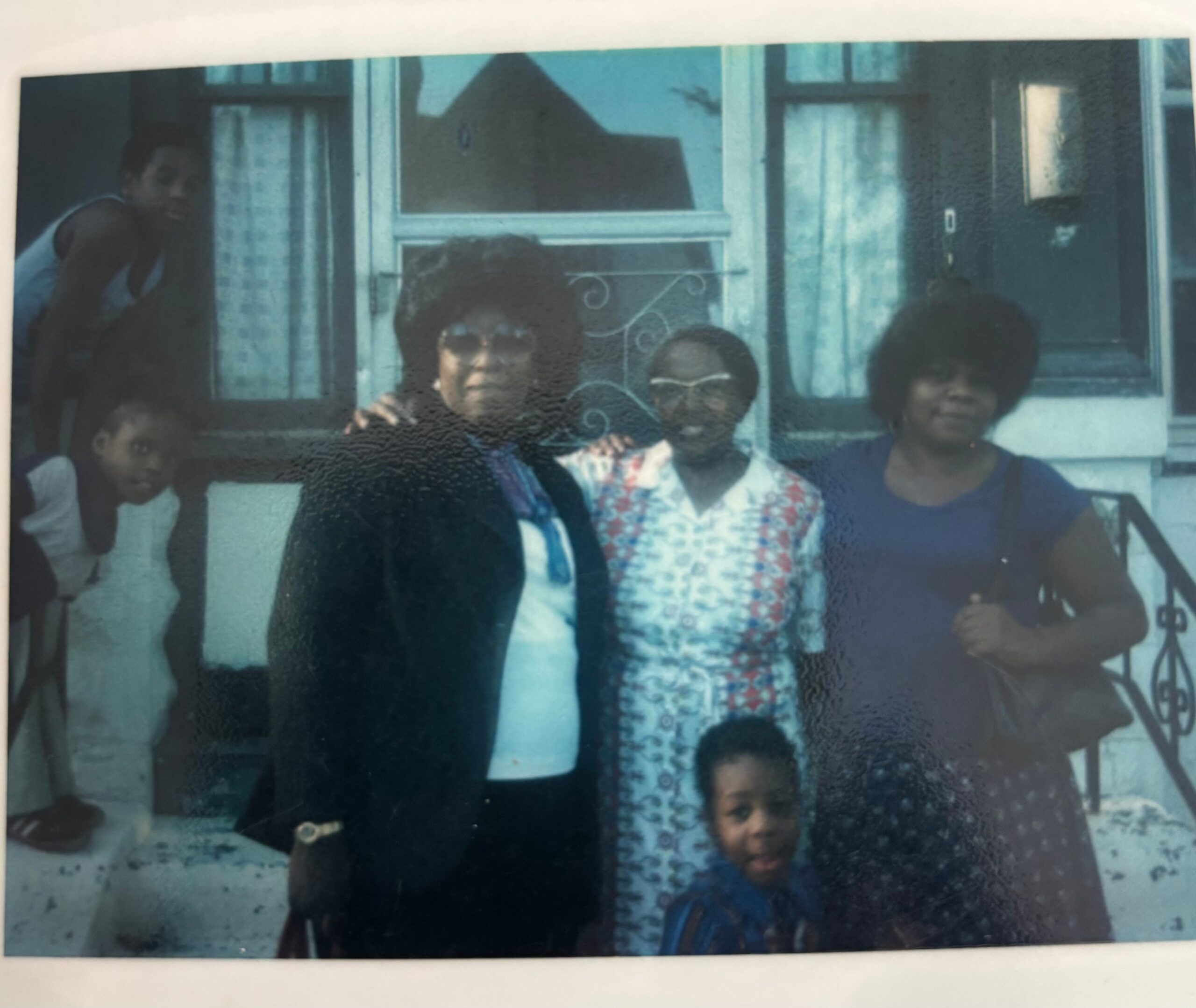 Group of five people (adults and children) posing outside a blue house in front of a doorway with a decorative railing.