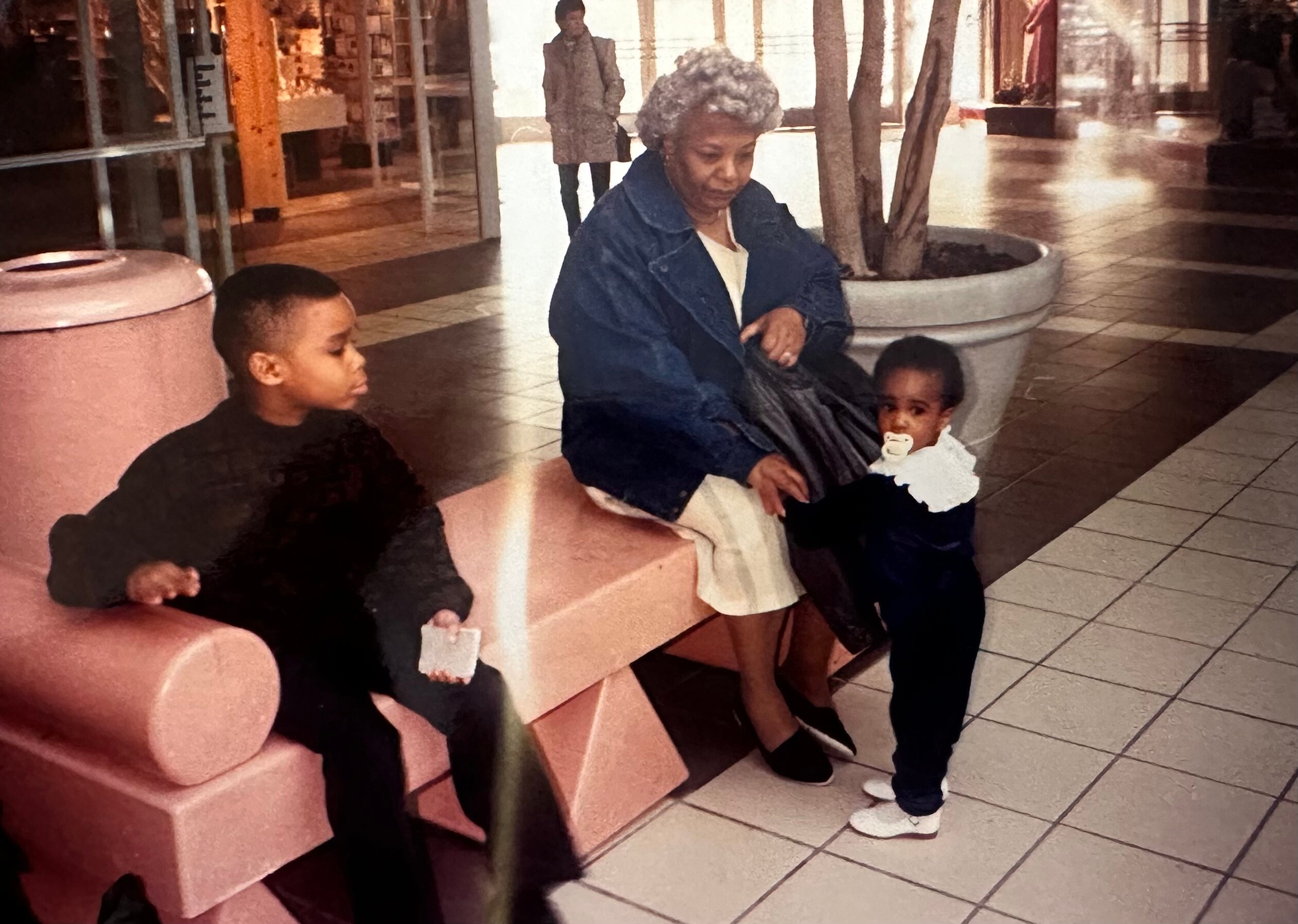 Elderly woman with gray hair in a blue jacket sits on a pink bench next to a young child with a pacifier in a shopping mall.”
