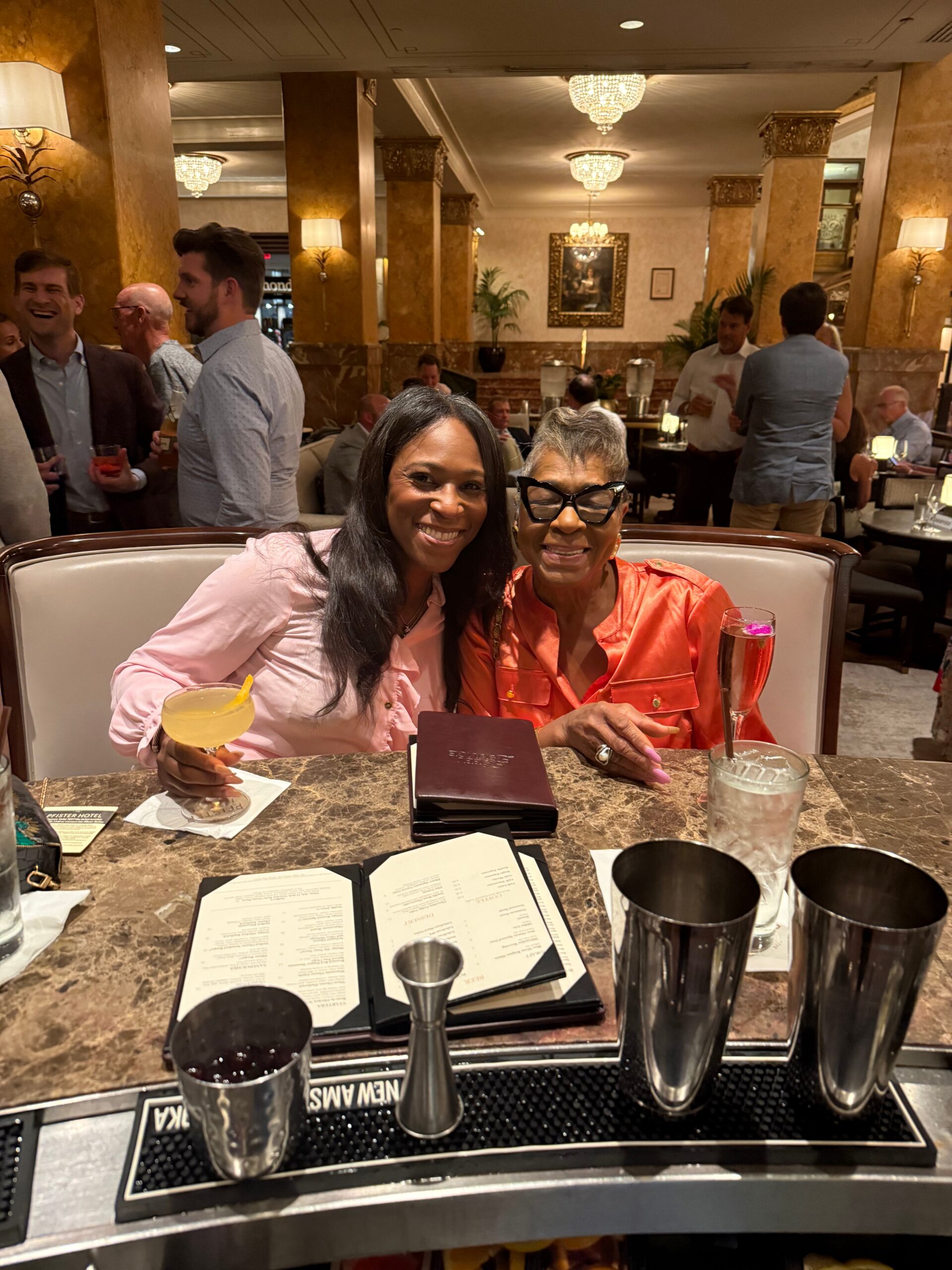 Two women smile at a restaurant table in a marble-top setting, with cocktails and menus visible.