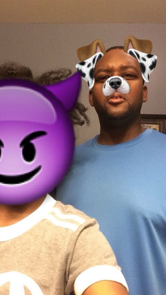 Two friends posing for a selfie; man in a blue shirt behind wears a dog-ear filter with nose, while the foreground person is obscured by a purple emoji face.