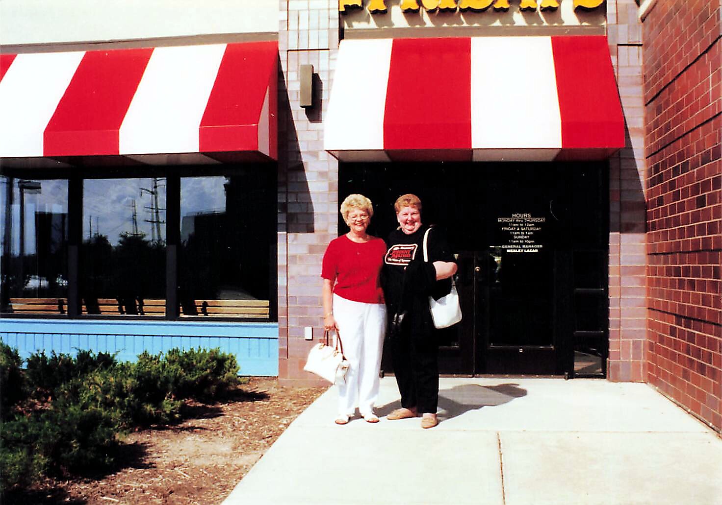 Two women stand together outside a storefront with red-and-white striped awnings, smiling at the camera.