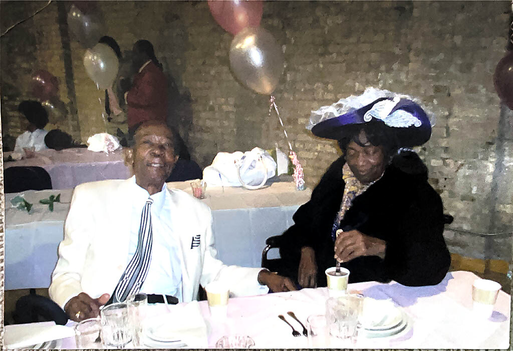 Two guests at a decorated banquet table: a smiling man in a white suit beside a woman wearing a large blue hat, balloons in the background.