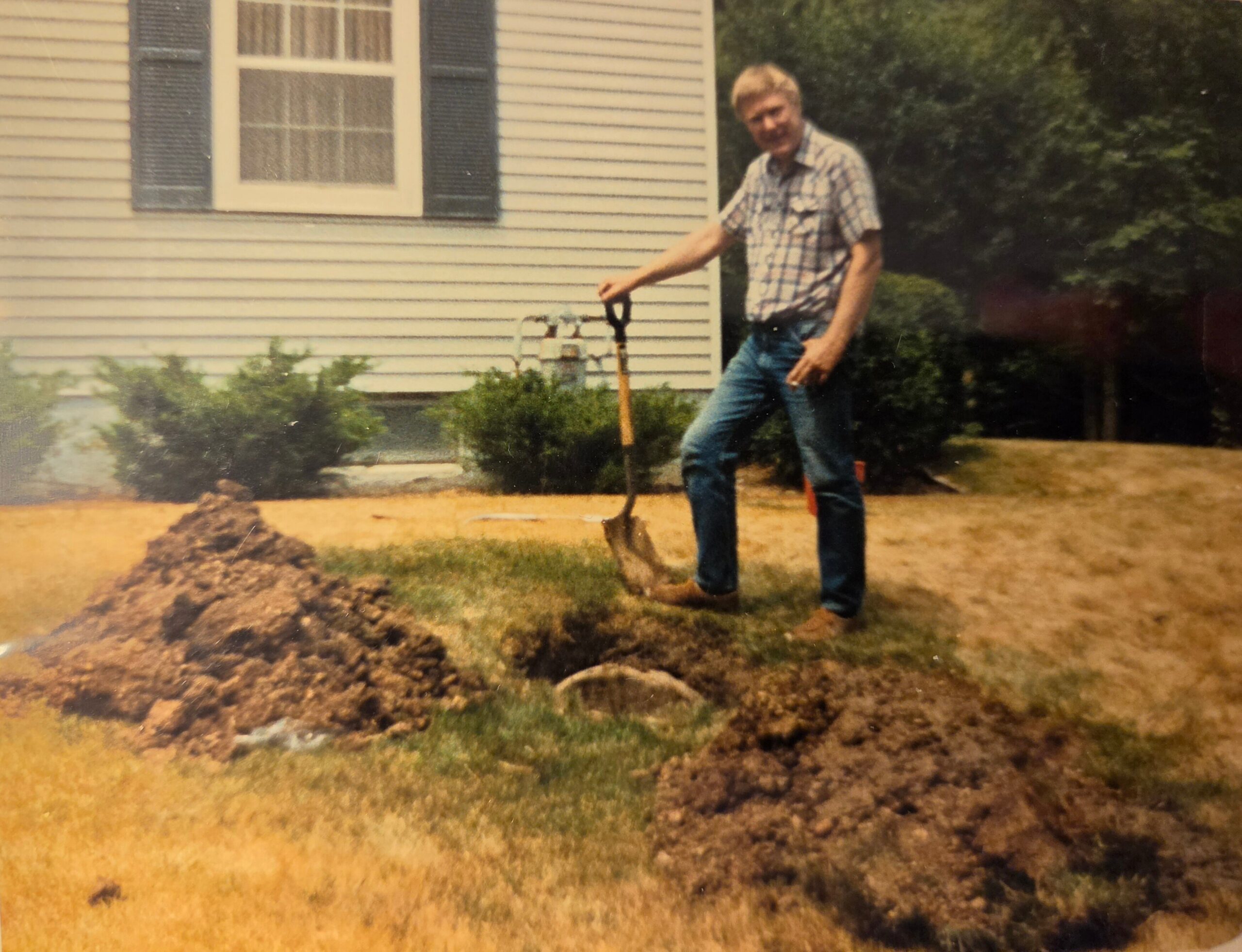 Man in a plaid shirt and jeans stands with a shovel in a dirt yard in front of a light siding house with dark shutters and a window behind him.